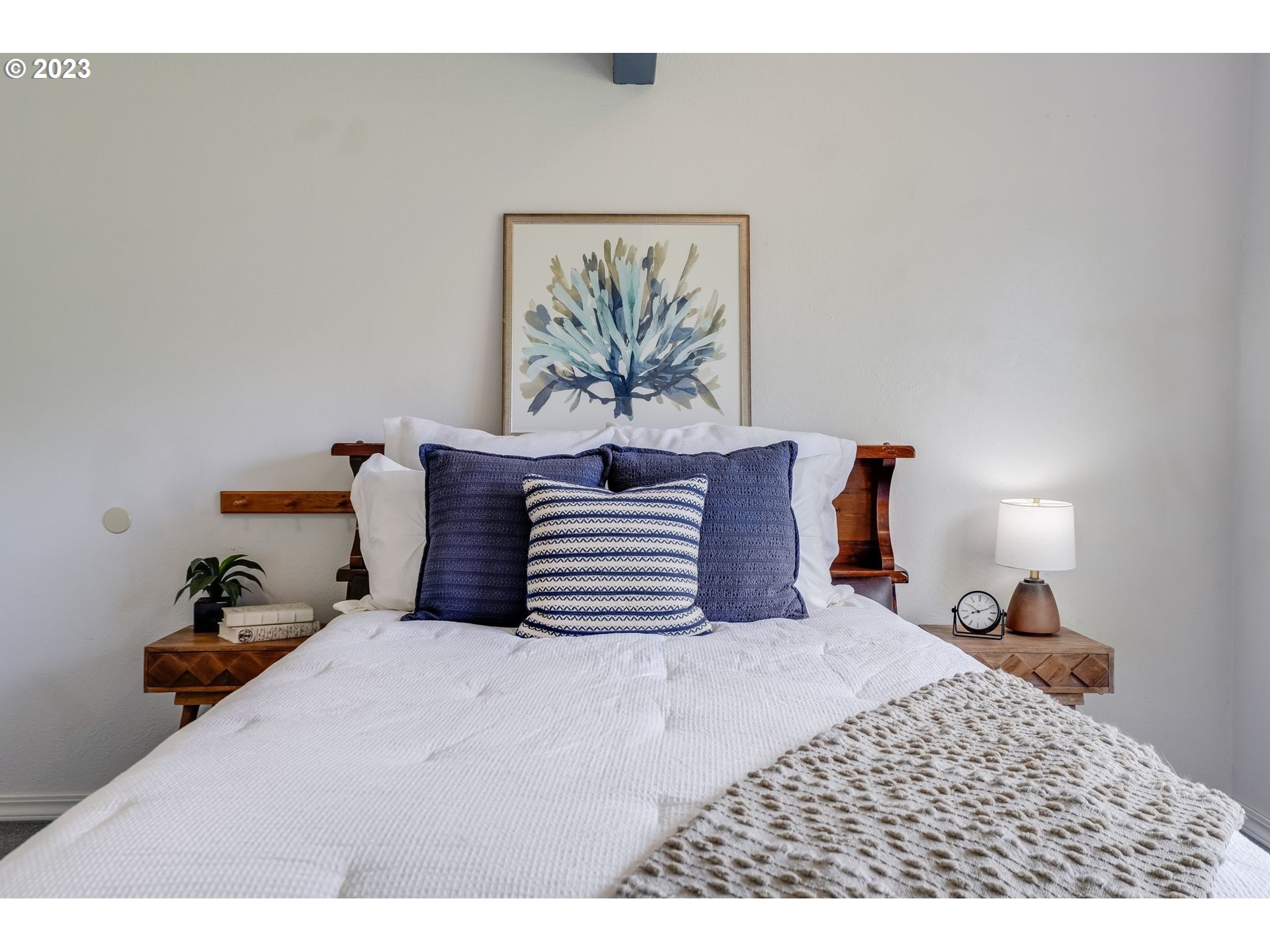 Staged bedroom with a white bed, navy pillows, wooden headboard, and framed blue botanical print.