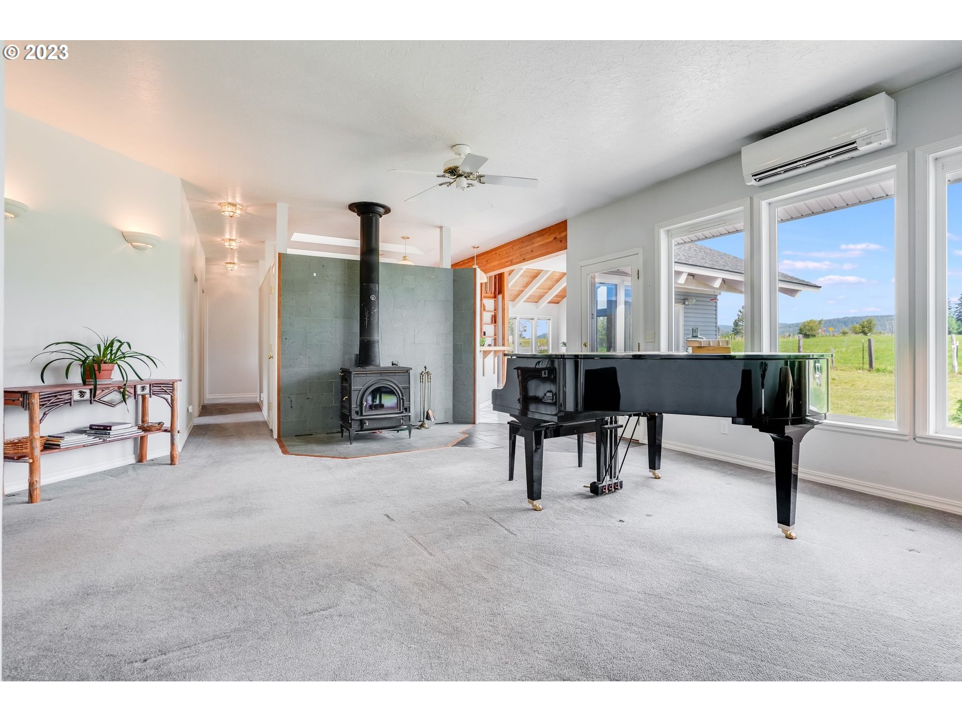 Living room with a black grand piano, tiled wood-stove hearth, and large windows overlooking a field.
