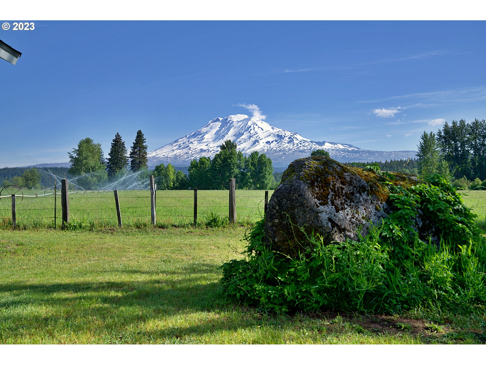 Grassy property with a fenced field and a large boulder, looking toward snow-covered Mount Adams.