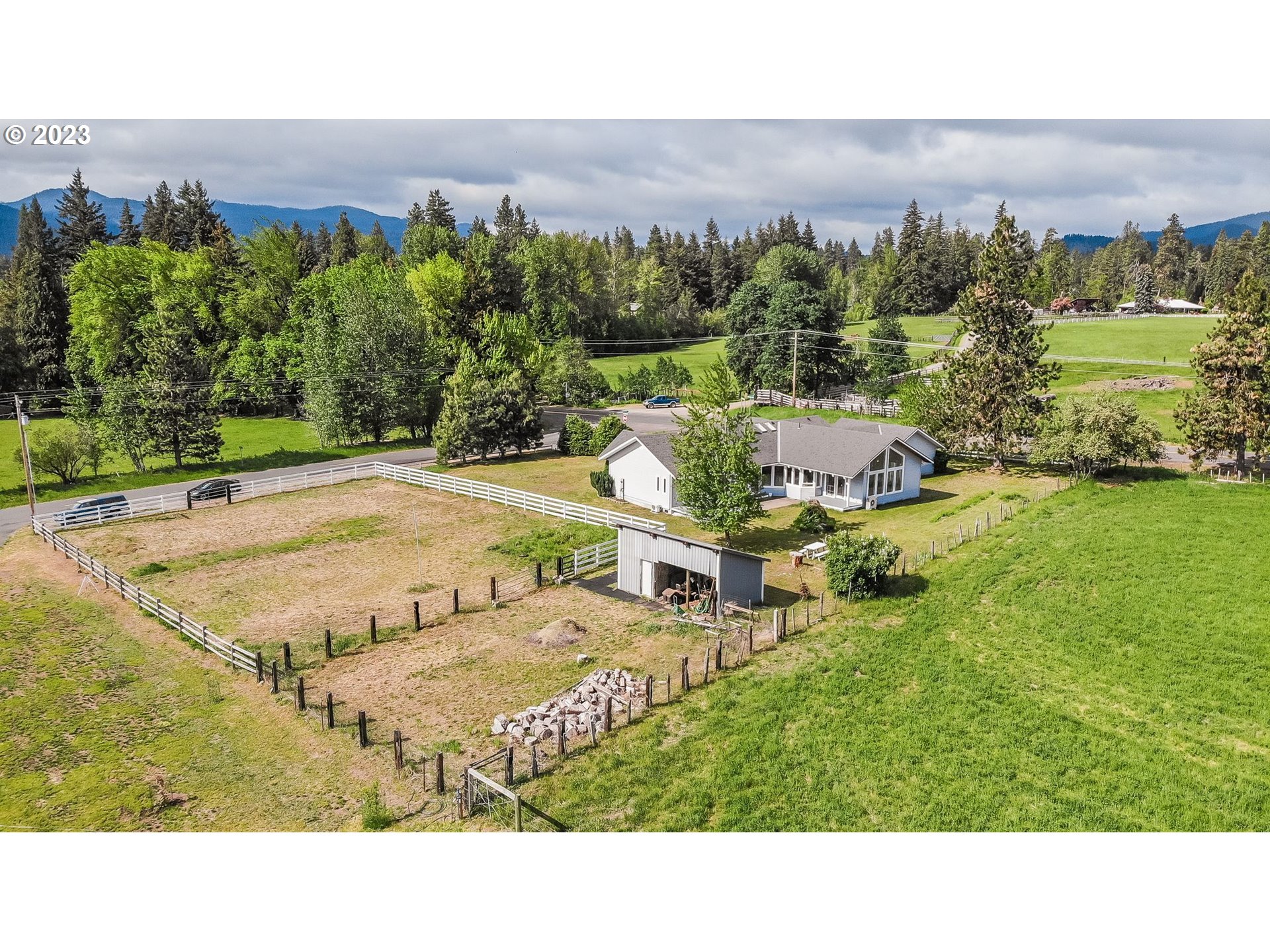 Aerial view of a white house, outbuilding, and fenced acreage with distant mountains.