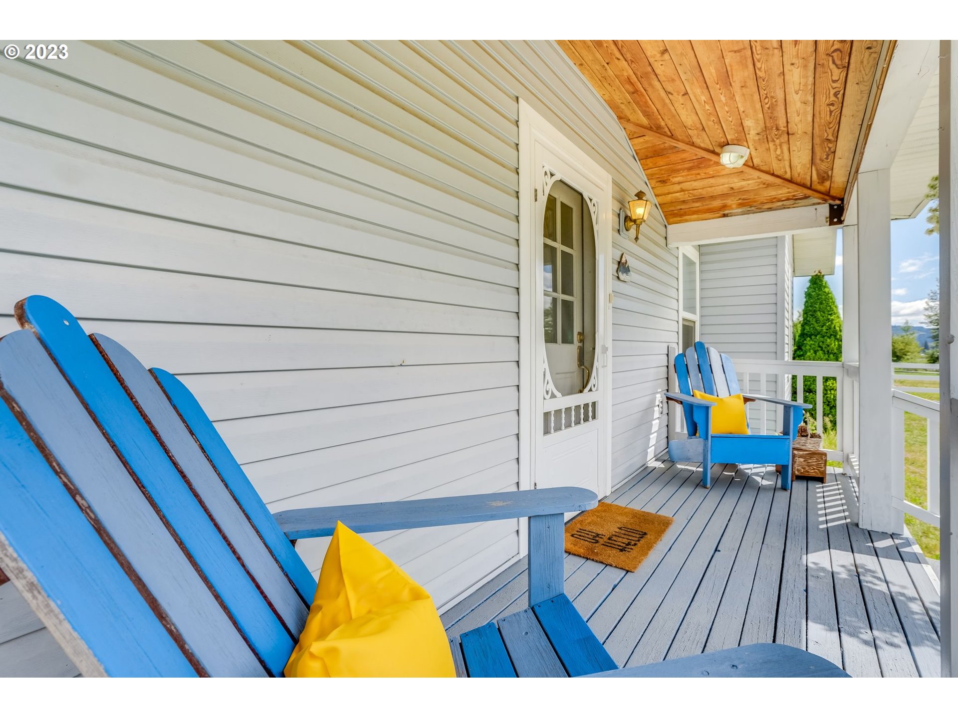 Covered porch with blue Adirondack chairs, yellow cushions, and a wood-paneled ceiling.