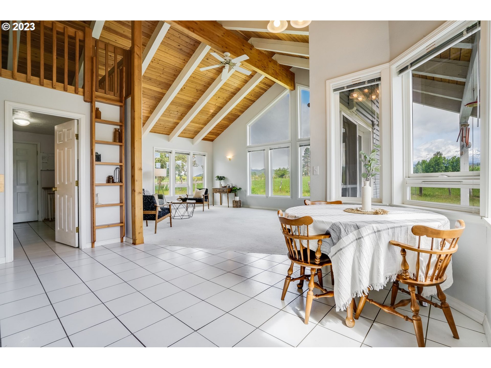 Open dining and living area with a vaulted wood ceiling, loft railing, and mountain views through large windows.