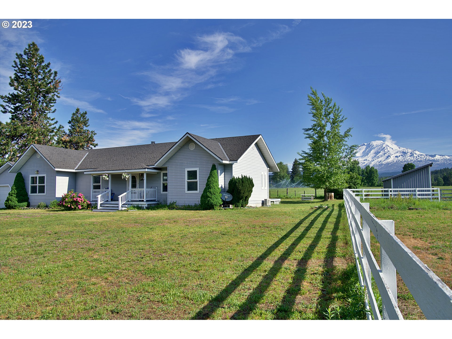 Gray single-story home with a covered porch, white fence, and snow-capped mountain view.