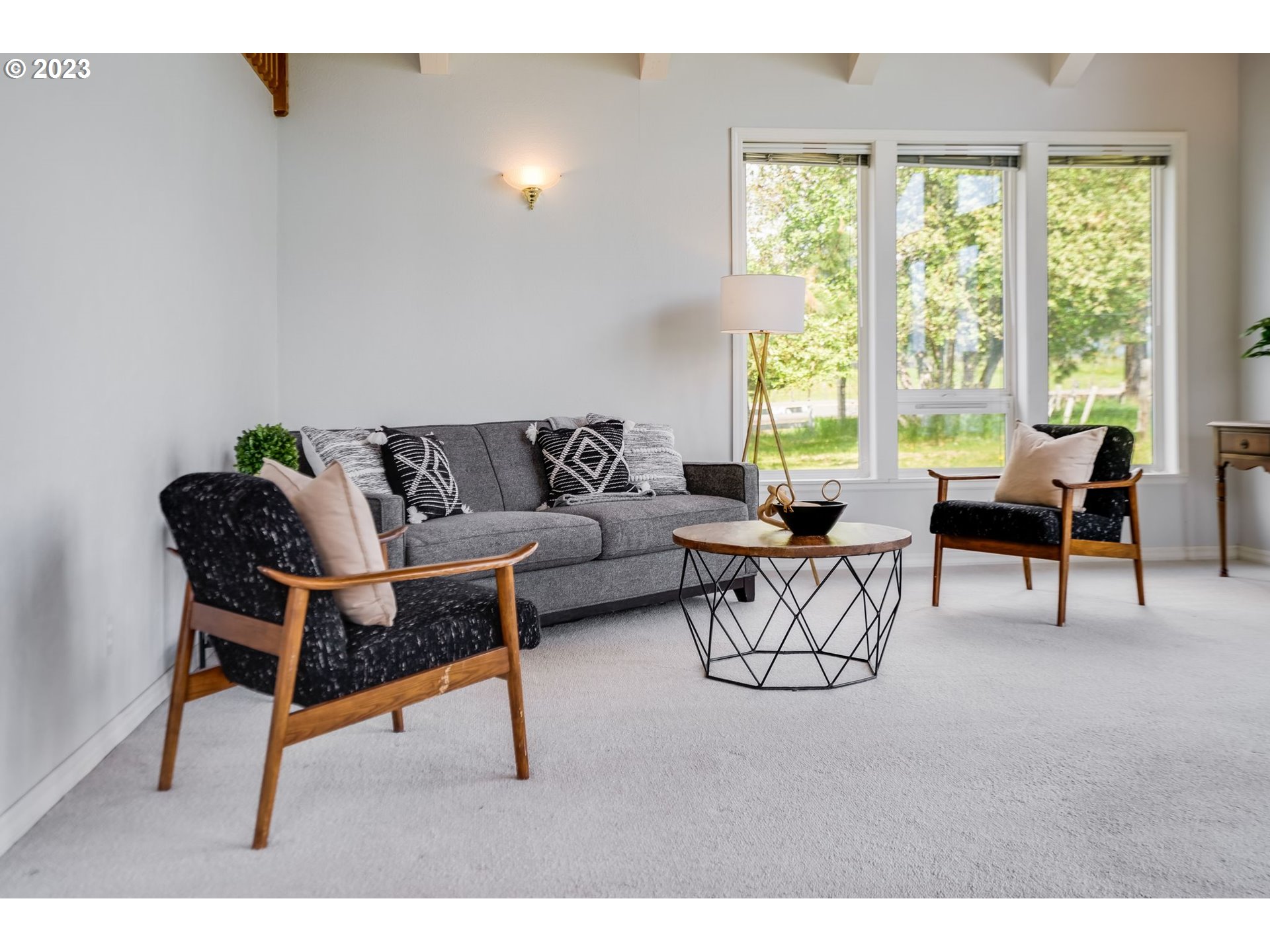 Living room with gray sofa, black-and-wood armchairs, and large windows overlooking a leafy yard.