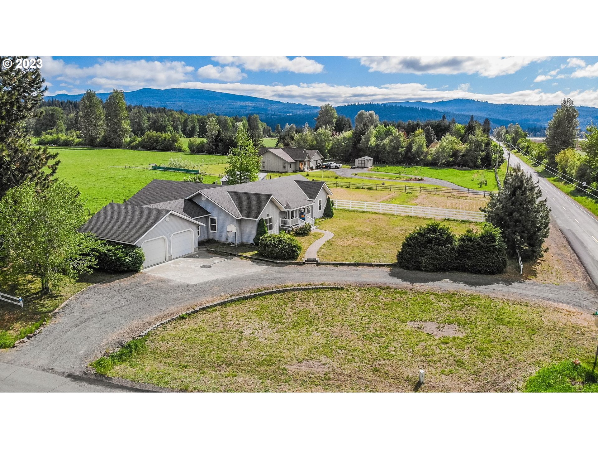Aerial view of a gray home with a two-car garage, broad lawn, and forested mountains beyond.