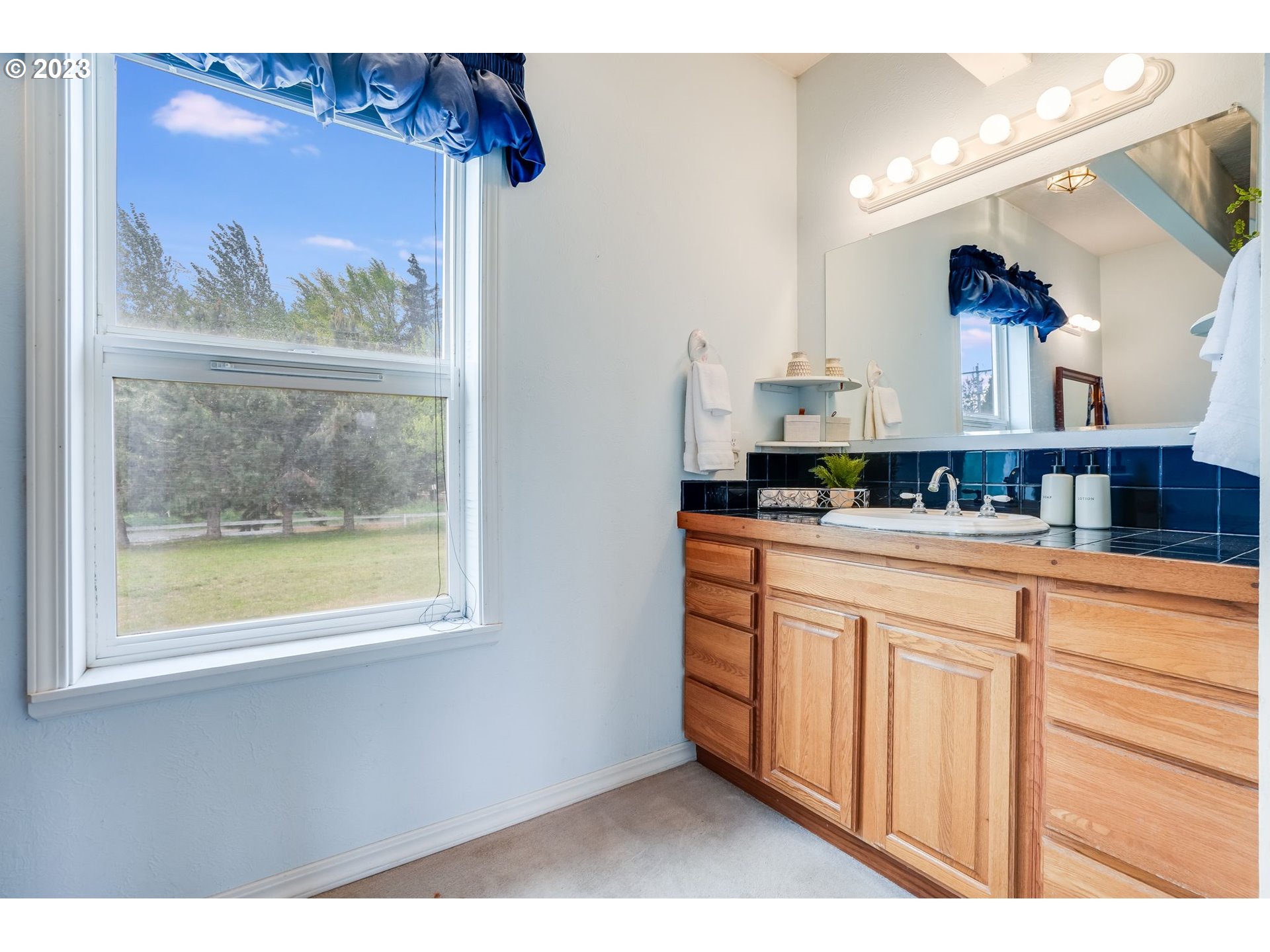 Bathroom with an oak vanity, blue tile, large mirror, and window overlooking a grassy yard.