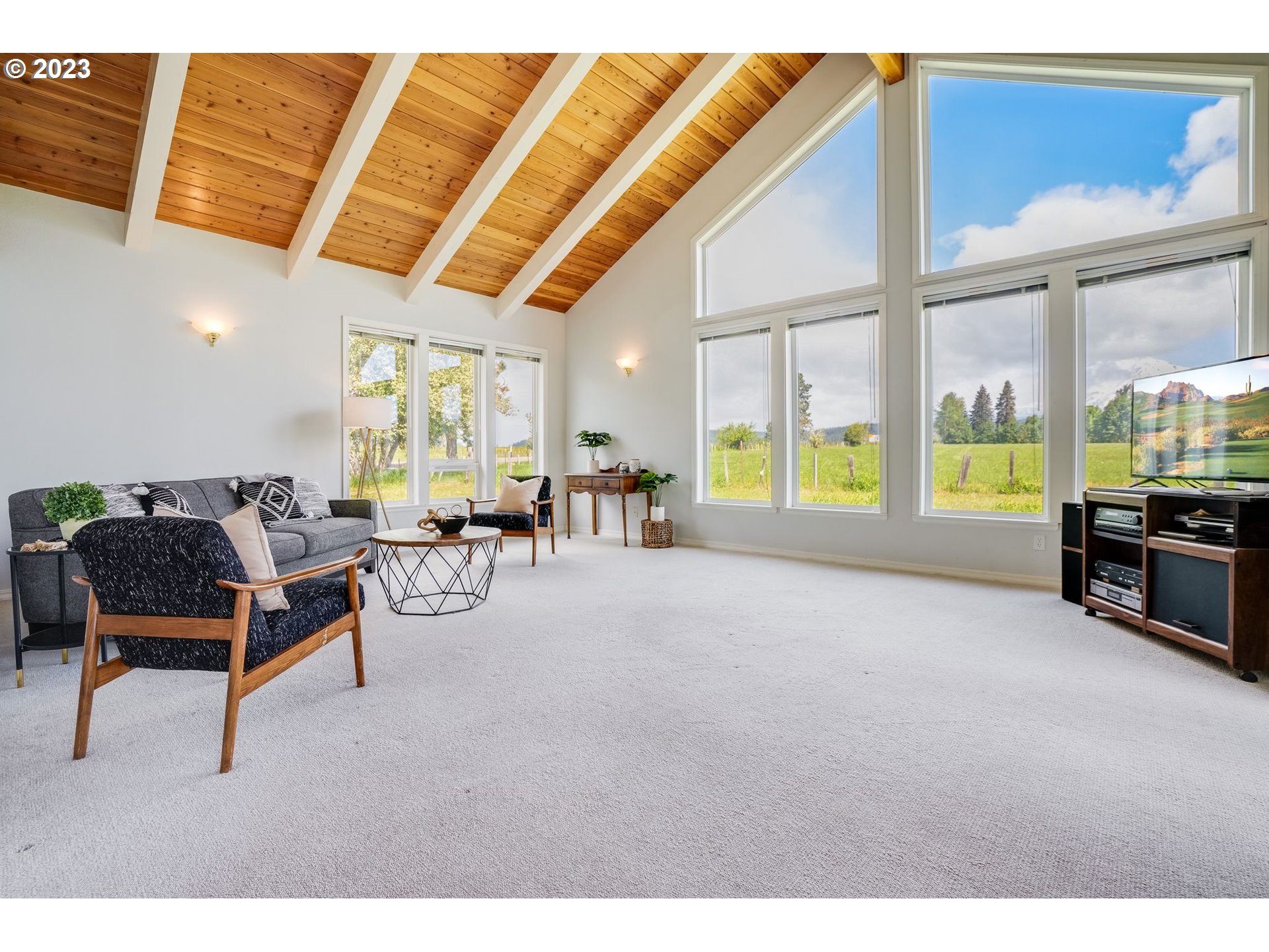 Living room with a vaulted wood ceiling, large windows, and views of fields and distant mountains.