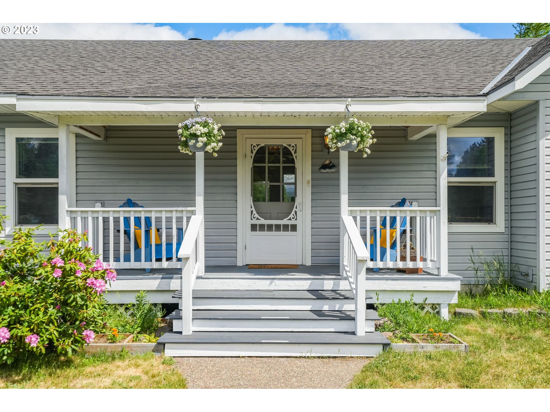 Gray house entrance with a covered porch, hanging flower baskets, and colorful Adirondack chairs.