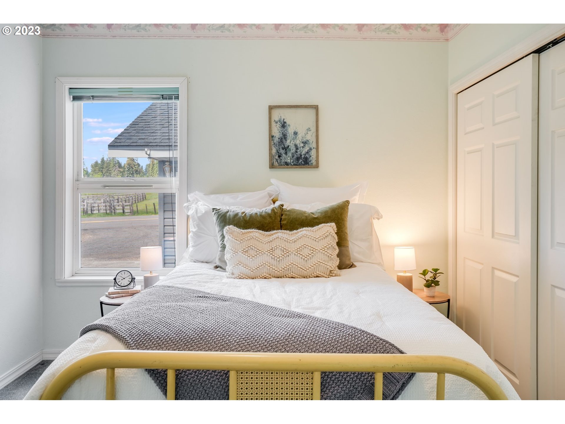 Staged bedroom with a white bed, olive accent pillows, gray throw, and a window overlooking the road.