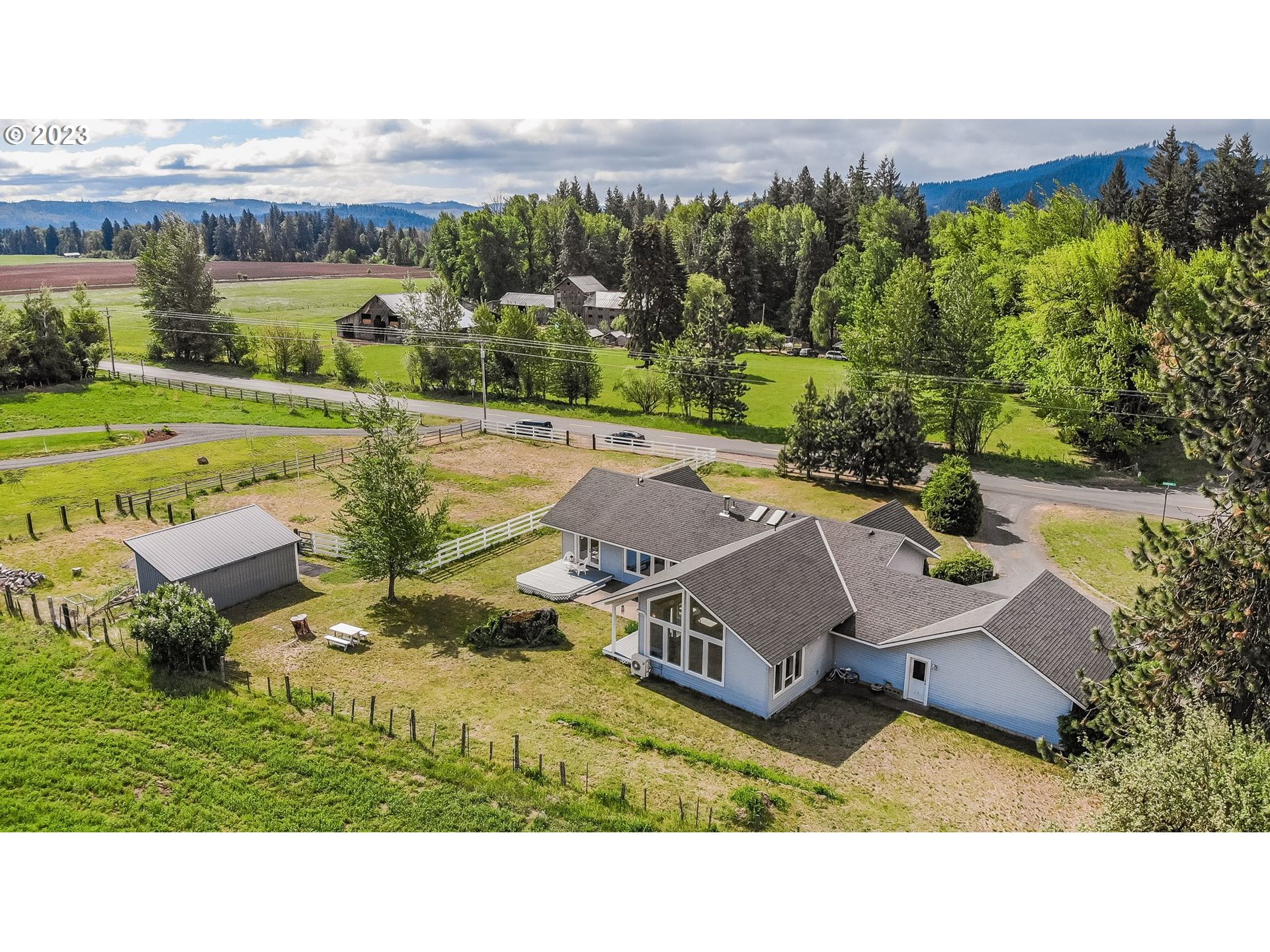 Aerial view of a rural house and fenced yard, with fields and wooded mountains beyond.