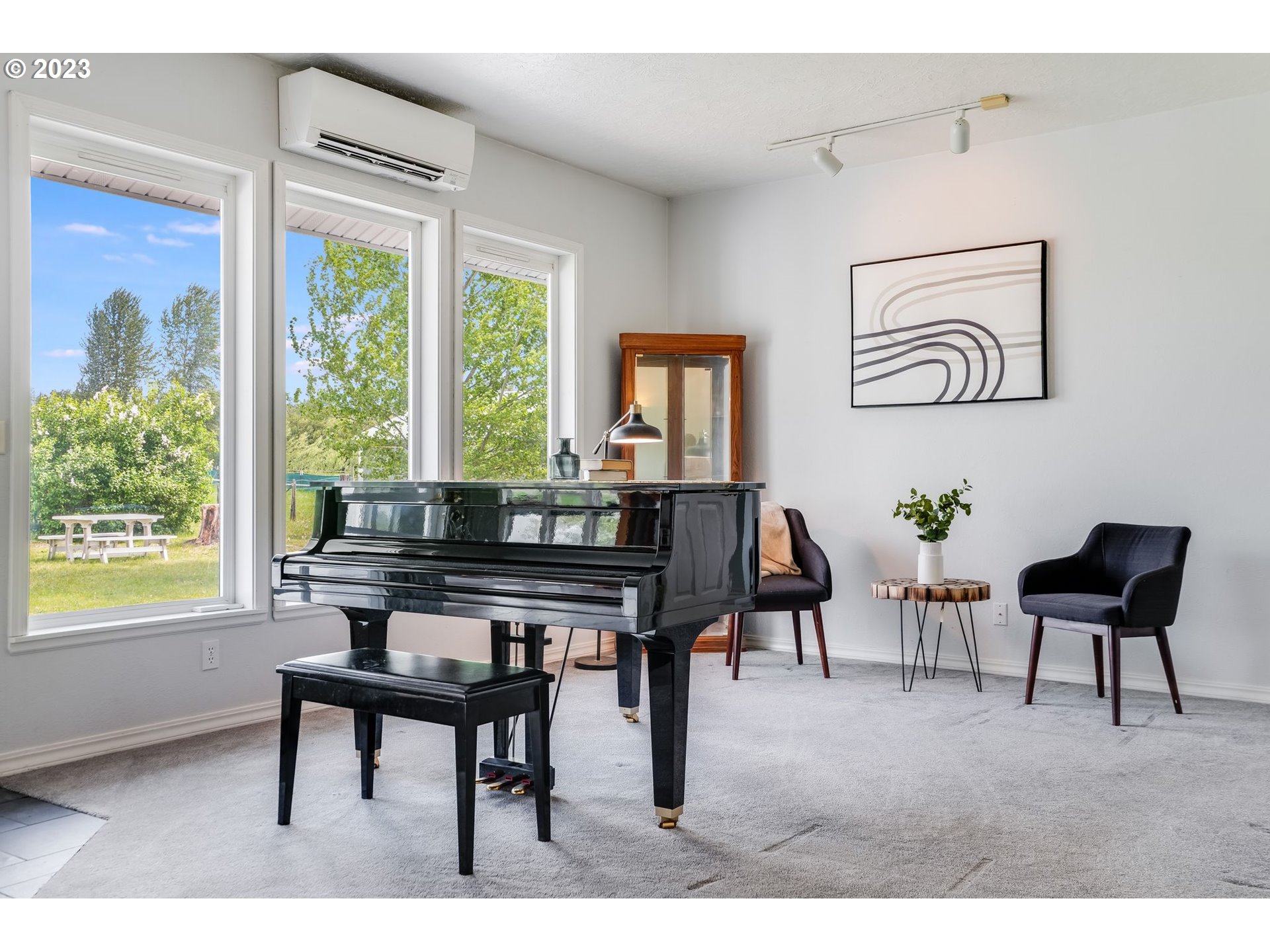 Music room with a black grand piano facing three tall windows overlooking leafy greenery.