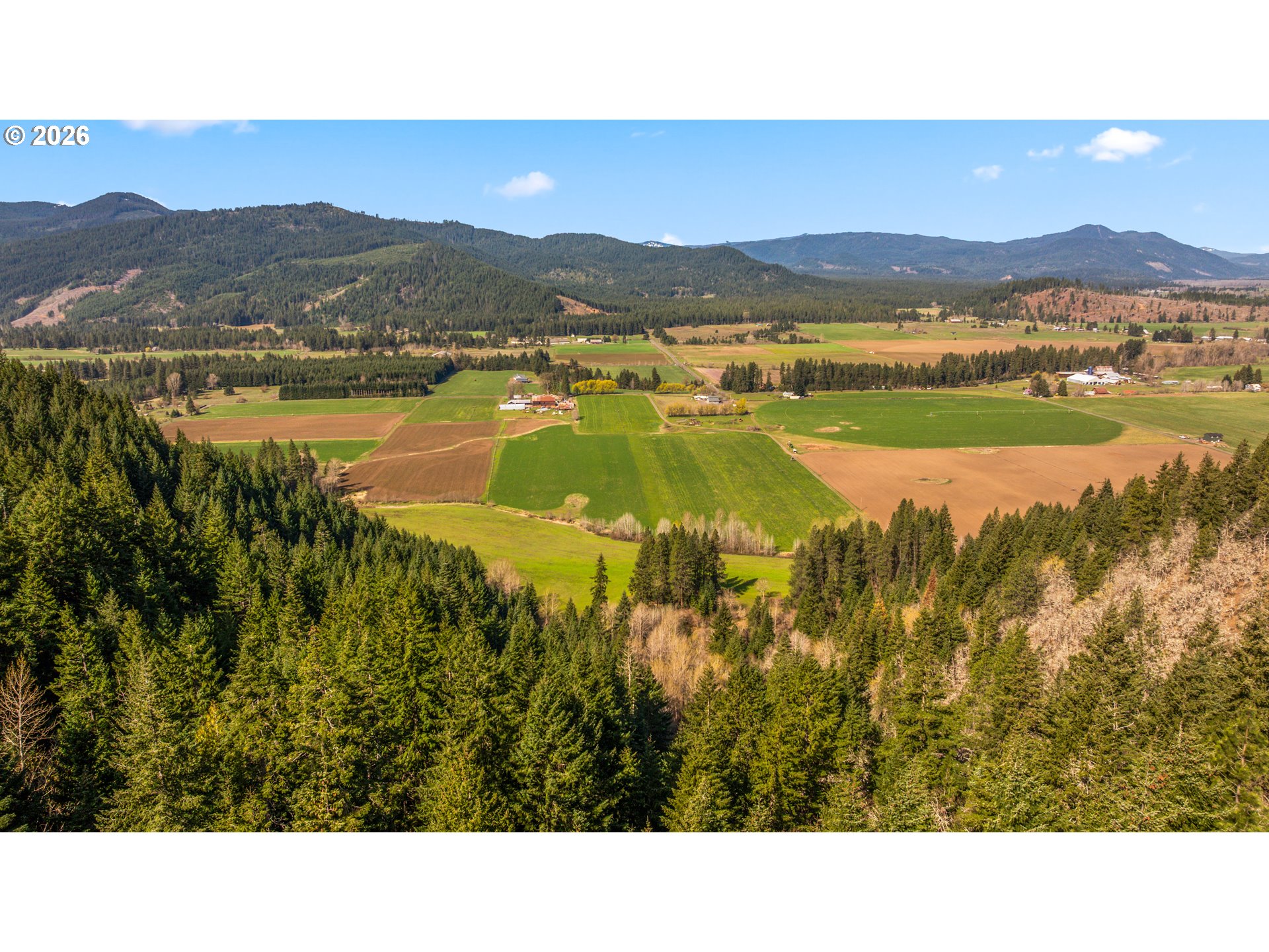 Aerial view of farm fields and rural buildings framed by evergreen forest, with mountain ridges beyond.