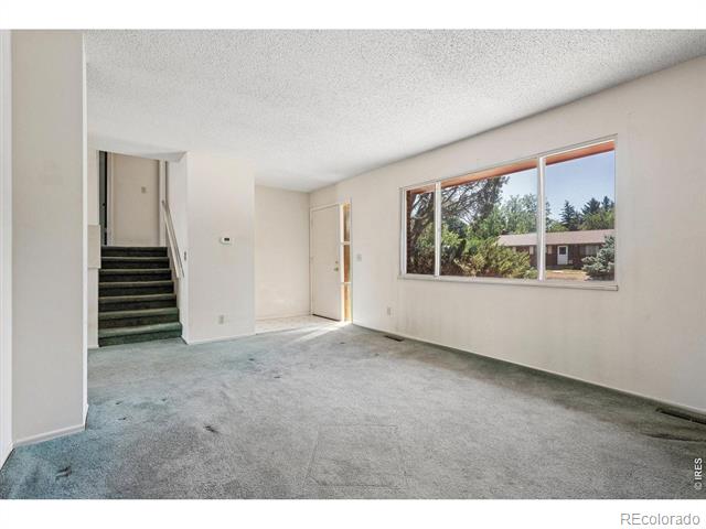 Empty living room with gray carpet, a staircase, and a wide window overlooking trees.