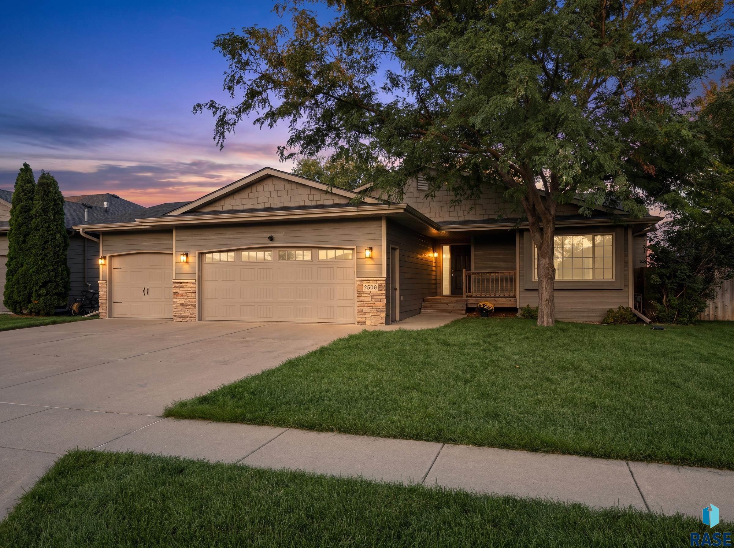 Single-story home with two garage doors and a large shade tree at dusk.
