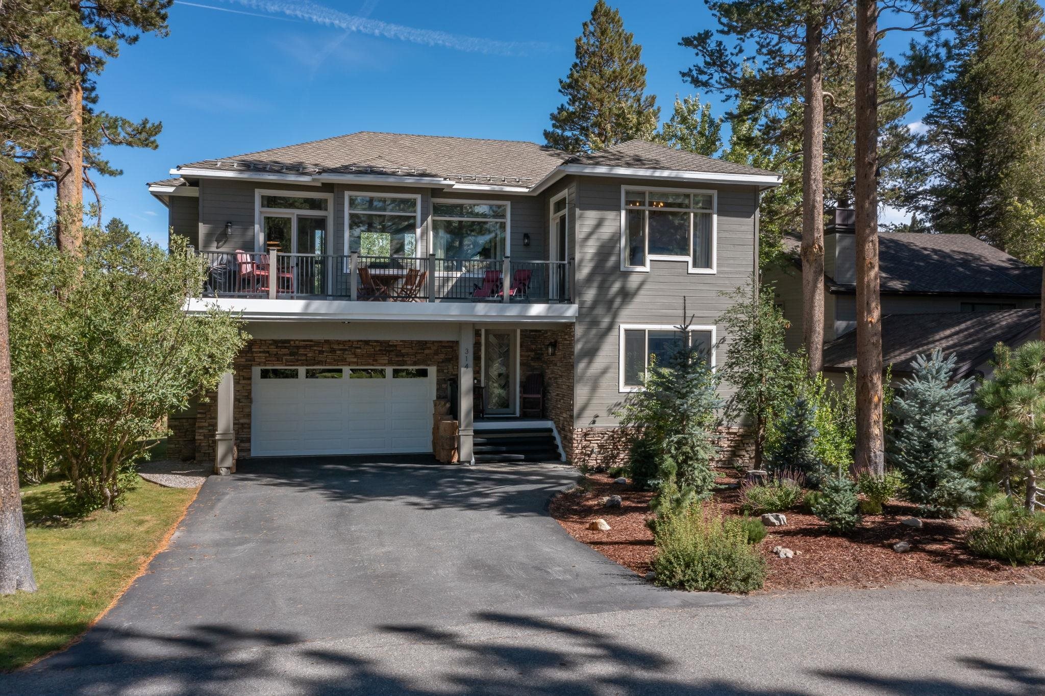 Two-story gray home with stone accents, a garage, and a furnished upper-level deck among pine trees.
