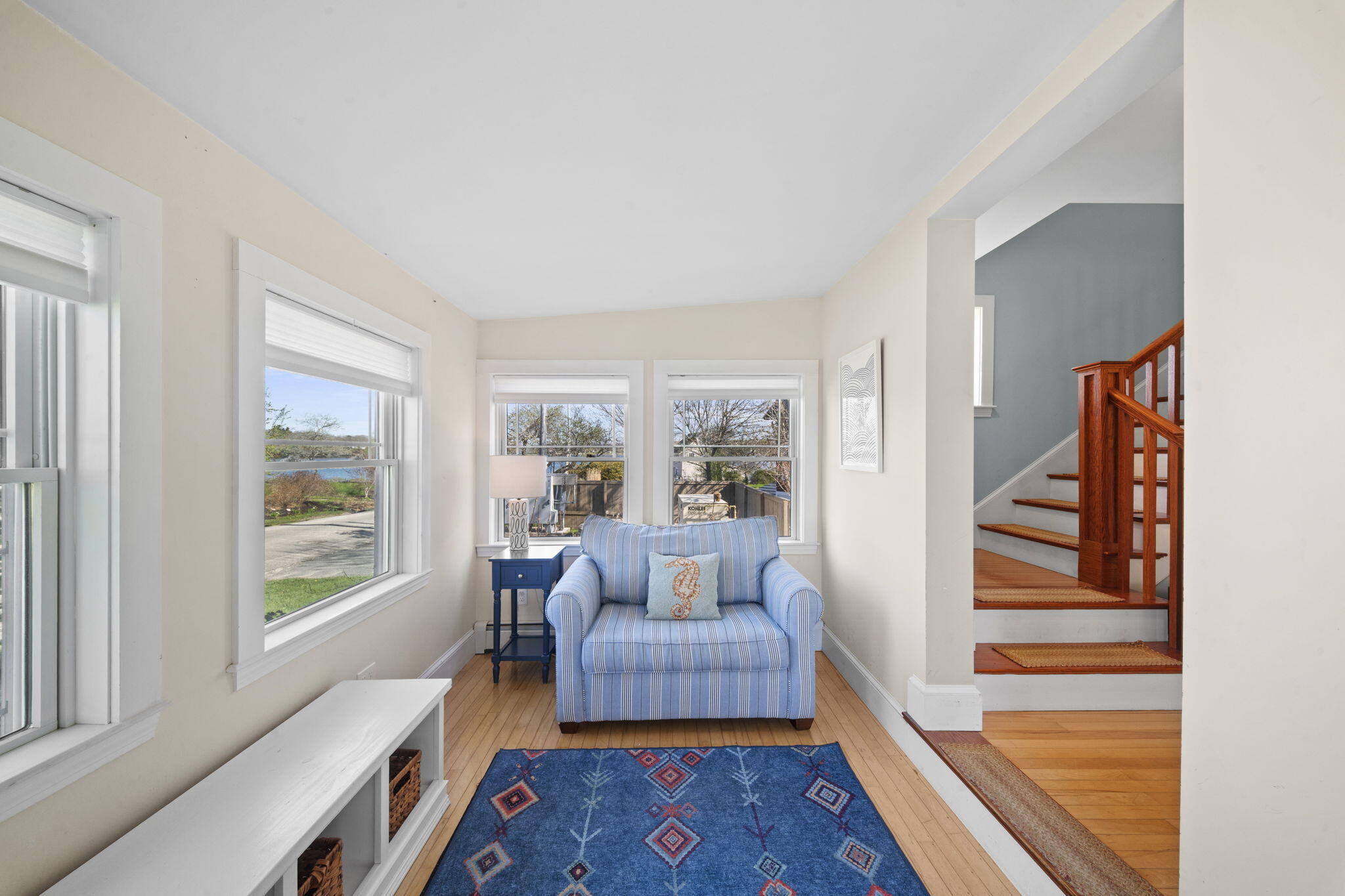 Sunlit sitting room with a blue striped armchair, large windows, and an open staircase.