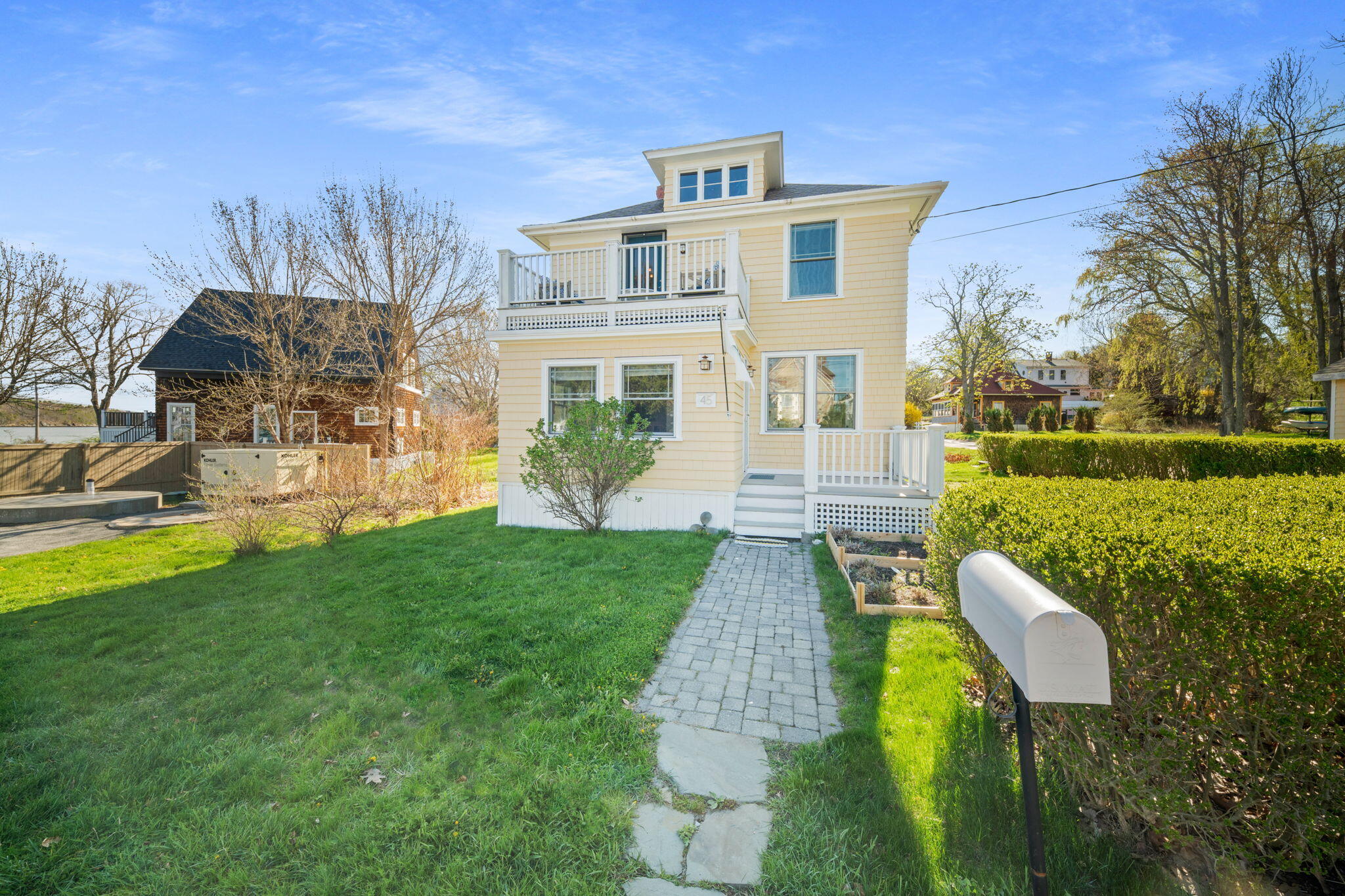 Pale yellow two-story house with white balconies, a paved walkway, and mailbox by the lawn.
