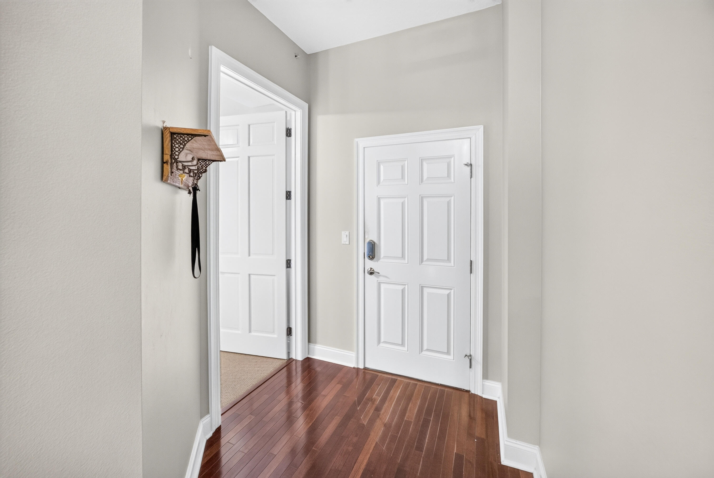 Entry foyer with a white paneled door, polished hardwood floors, and an open doorway to a carpeted room.