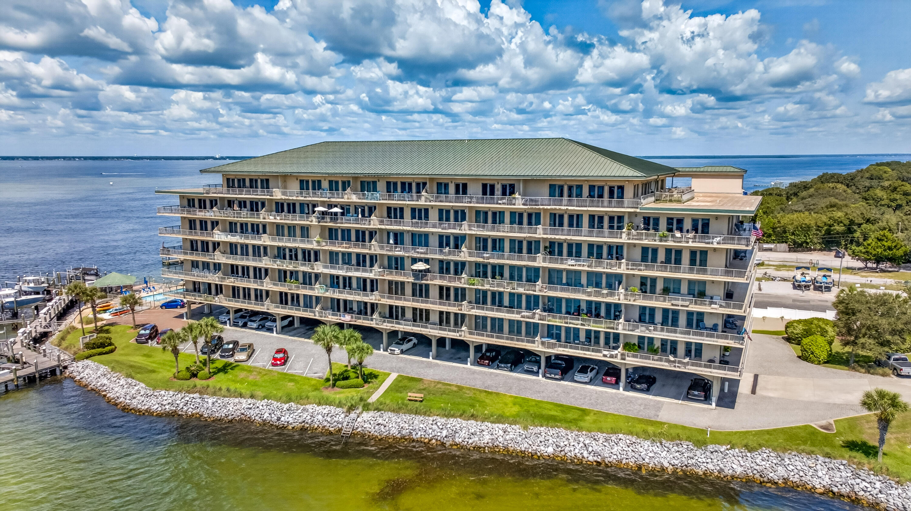 Waterfront condominium with rows of balconies and covered parking beside a rocky shoreline.