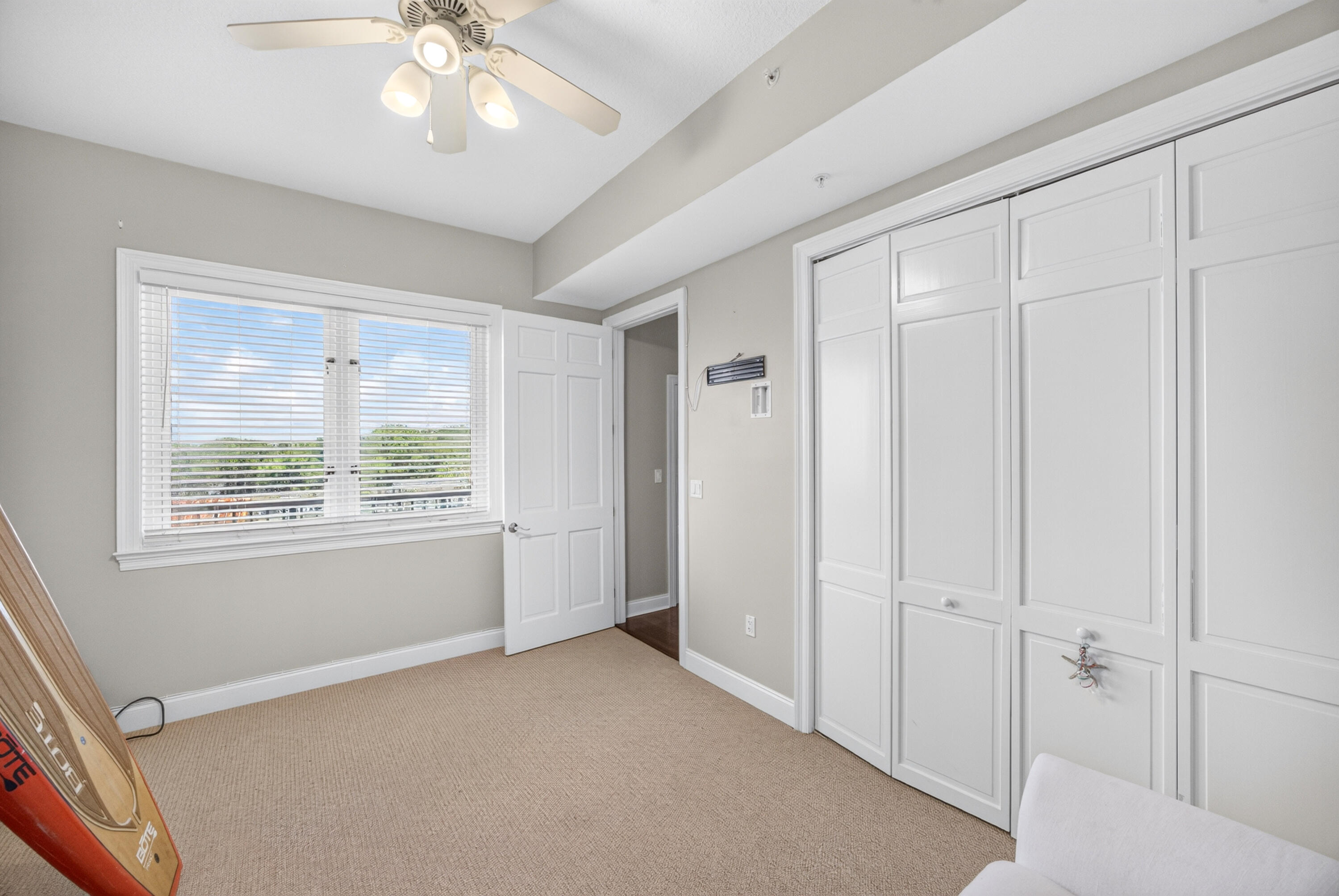 Empty carpeted room with white built-in closets, a ceiling fan, and surfboards leaning by the window.