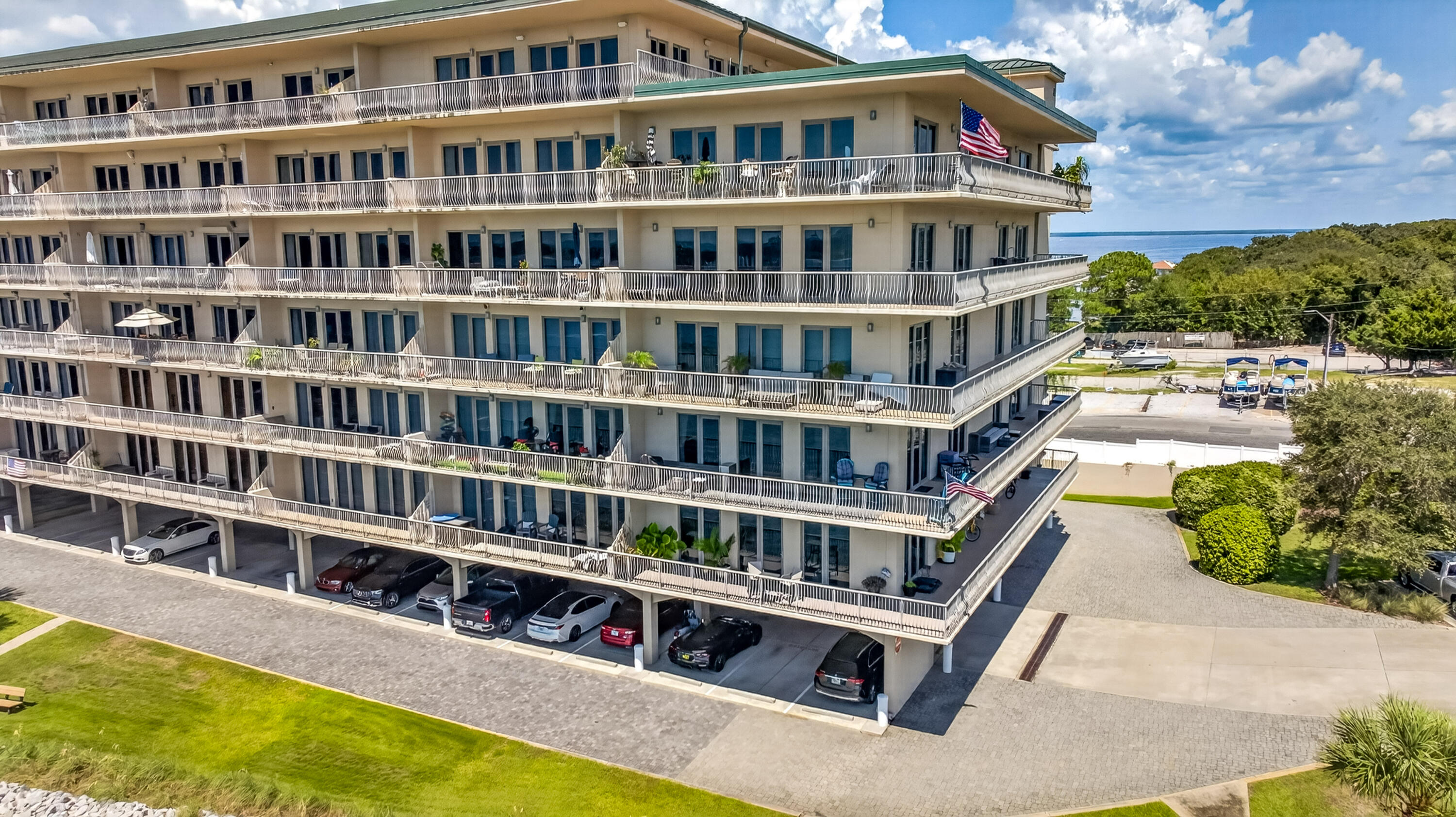 Multi-story condominium with wraparound balconies and covered parking, with coastal water visible beyond the trees.