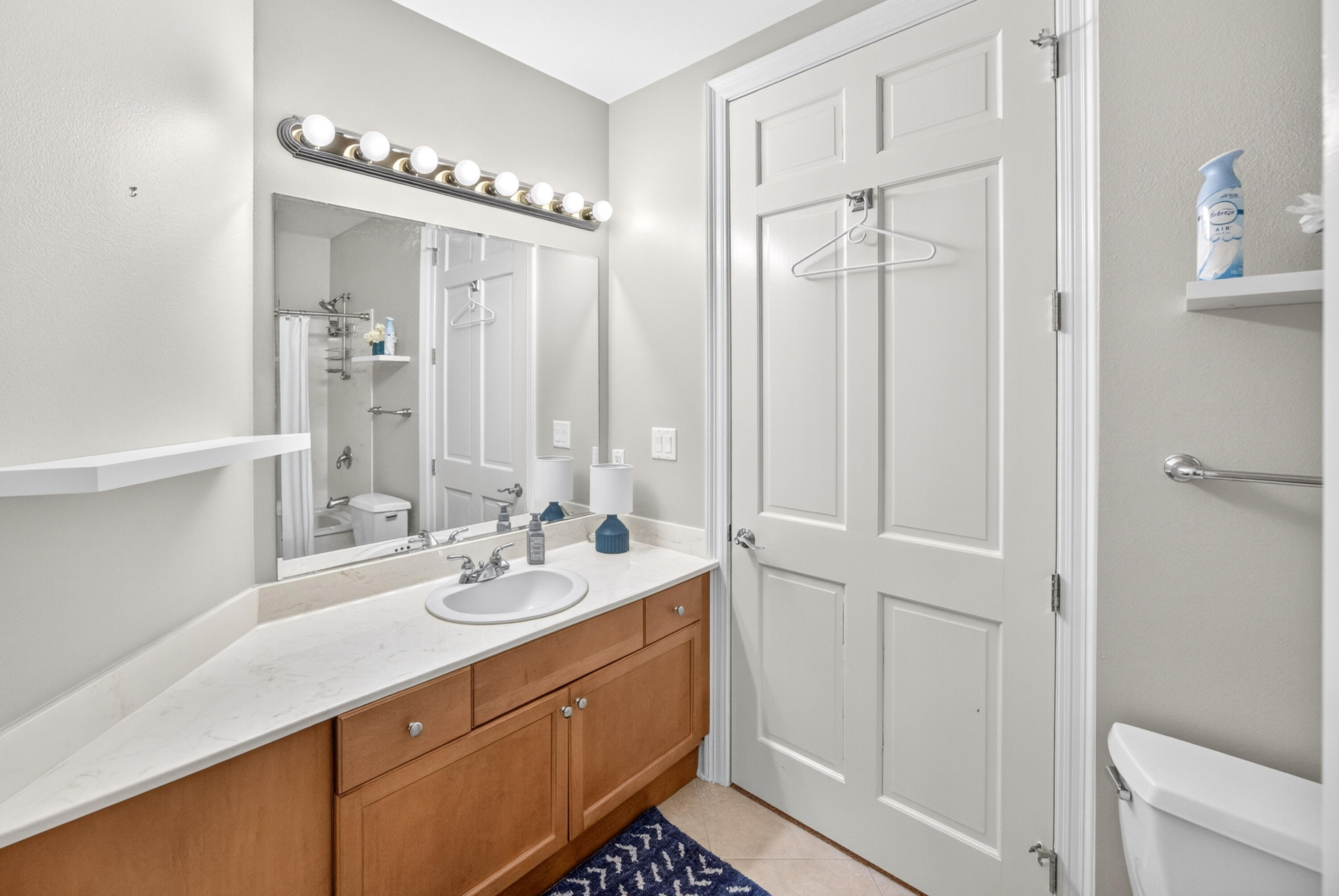 Bathroom with a long wood vanity, white countertop, large bulb-lit mirror, and shower reflected in the glass.