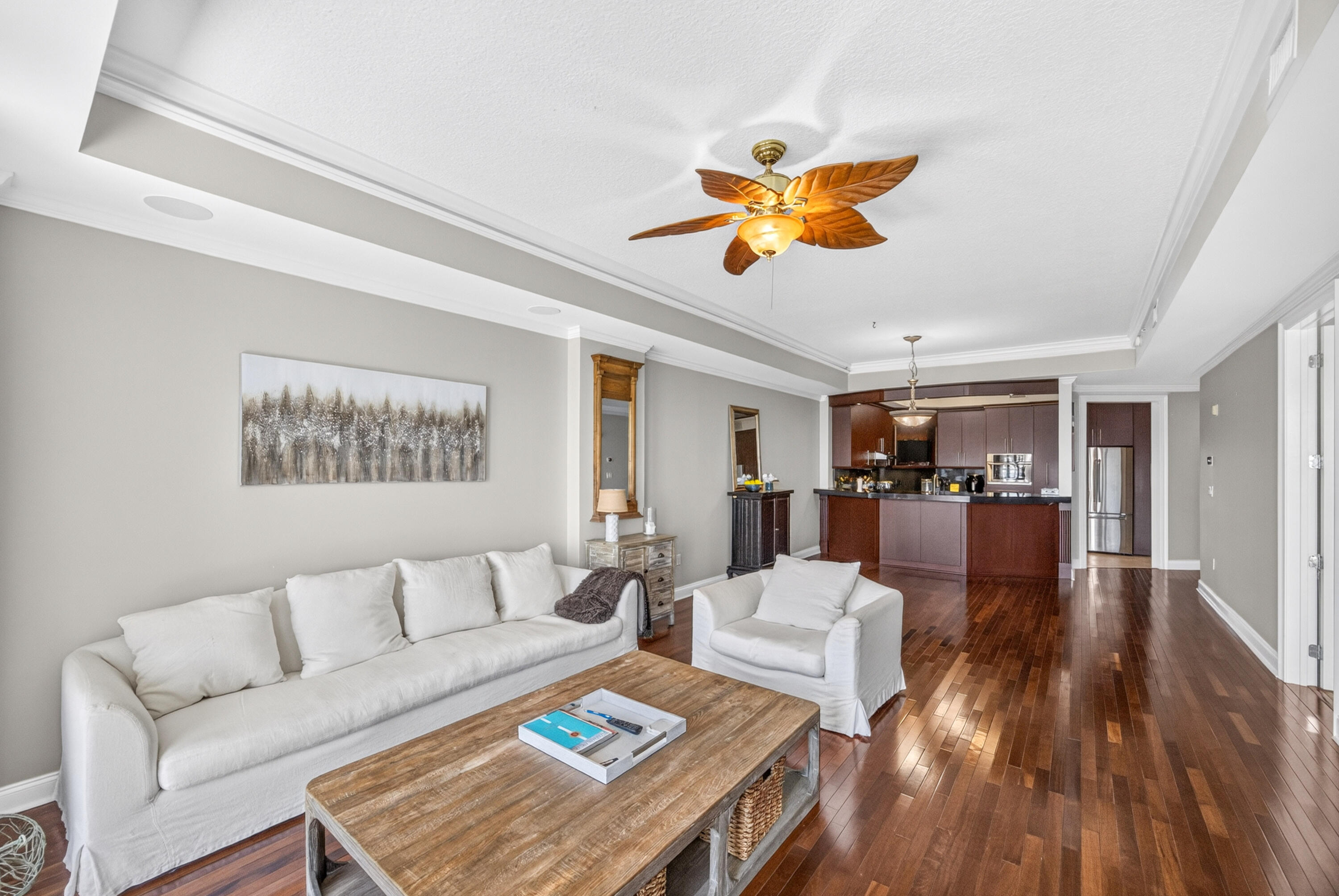 Open-plan living room with white seating, polished wood floors, and a leaf-blade ceiling fan beside the kitchen.