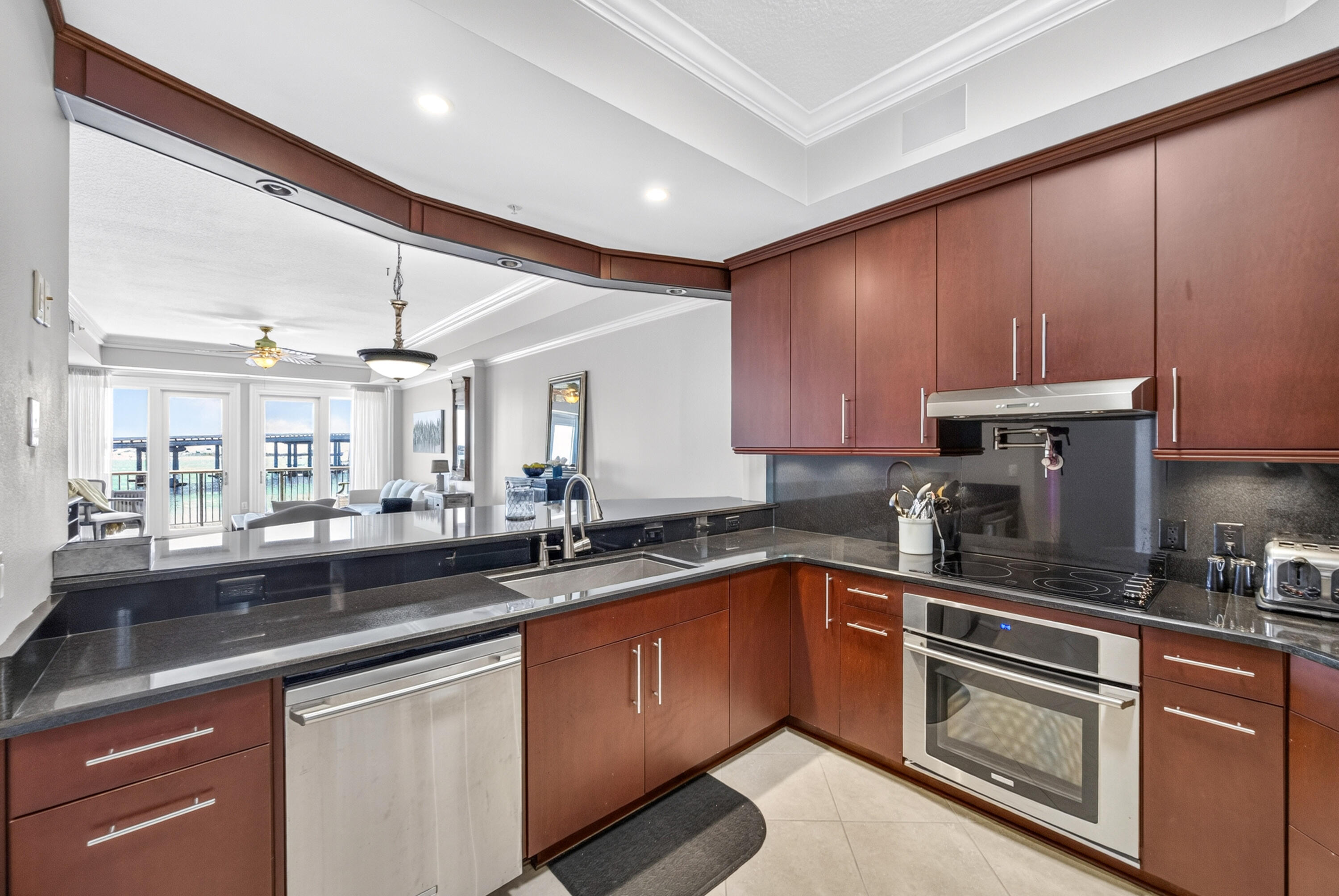 Condo kitchen with dark stone counters and reddish-brown cabinets, opening to a living room with water views.