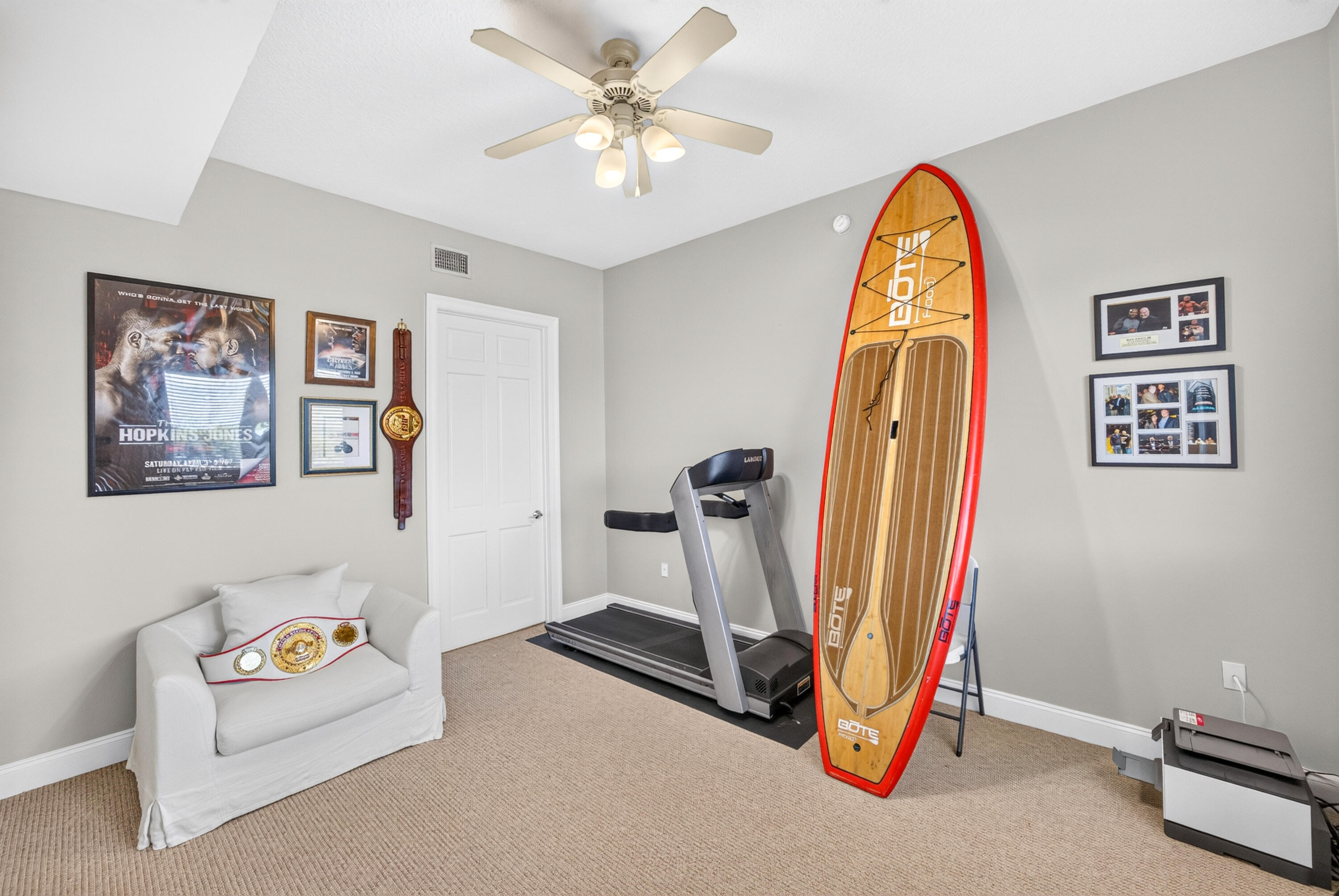 Exercise room with a treadmill, upright paddleboard, and championship belts displayed on a white chair.