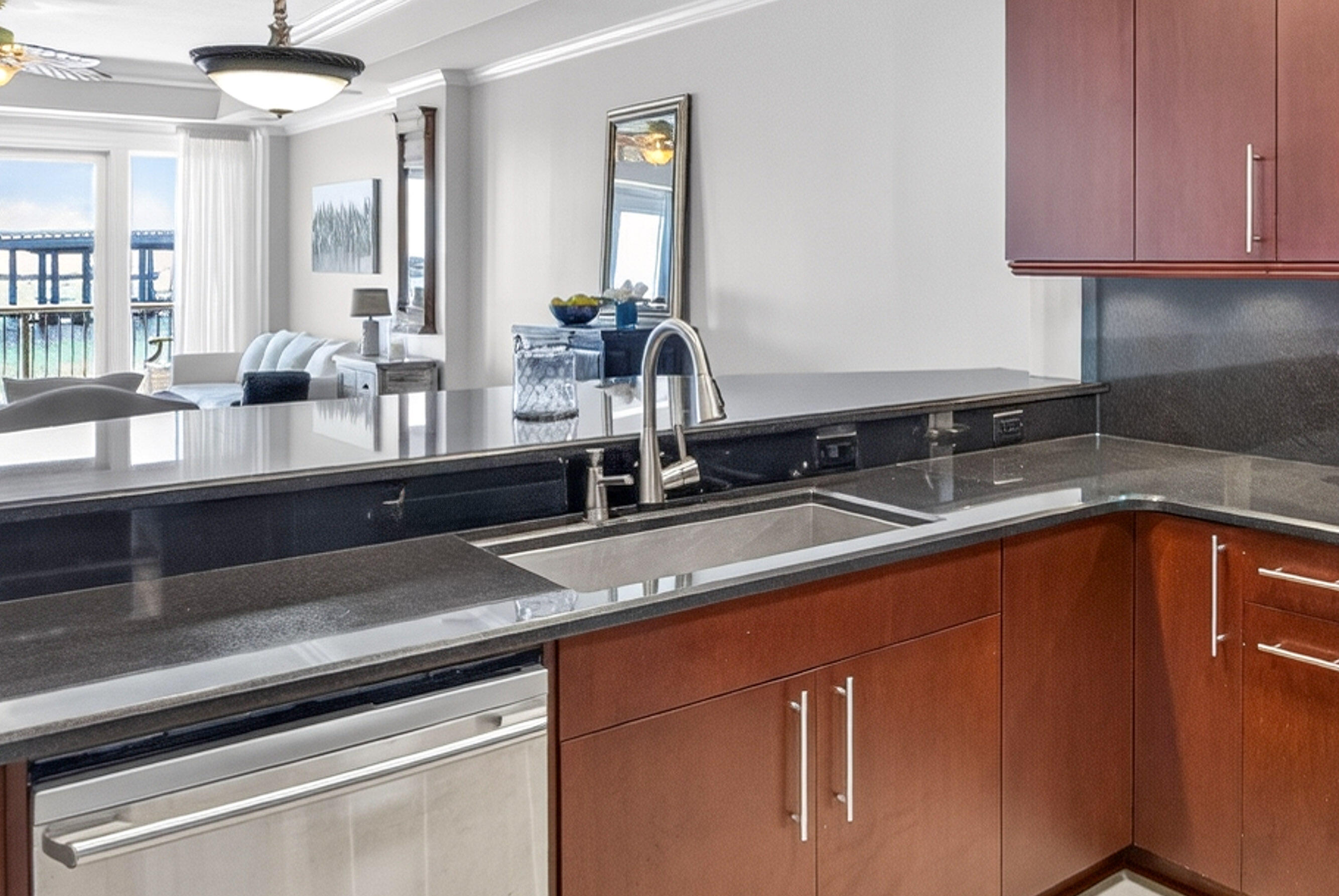 Condo kitchen with reddish-brown cabinets and dark counters, opening to a living area and balcony water views.