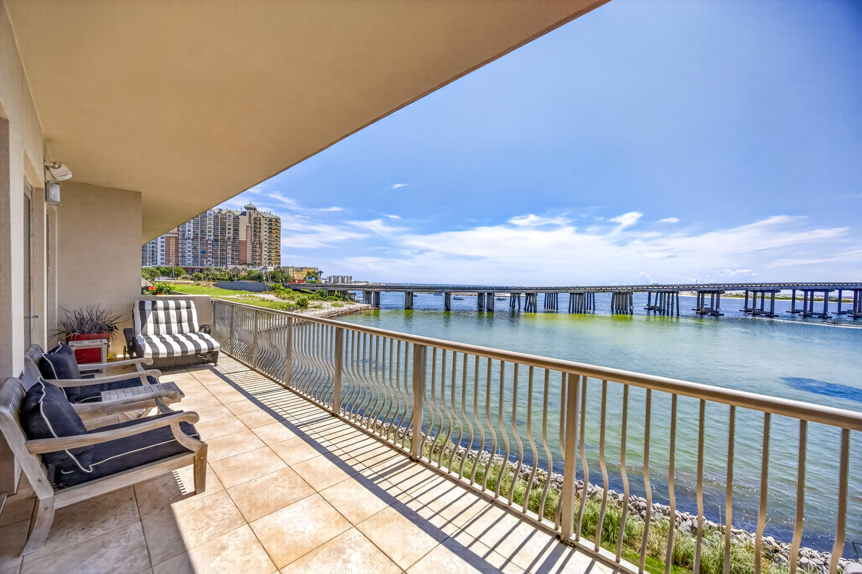 Waterfront balcony with cushioned chairs overlooking the water, bridge, and high-rise buildings.