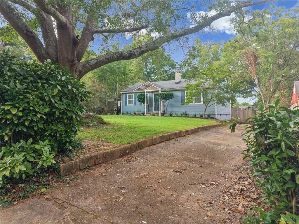 Blue single-story house beyond a driveway, framed by mature trees and shrubs.