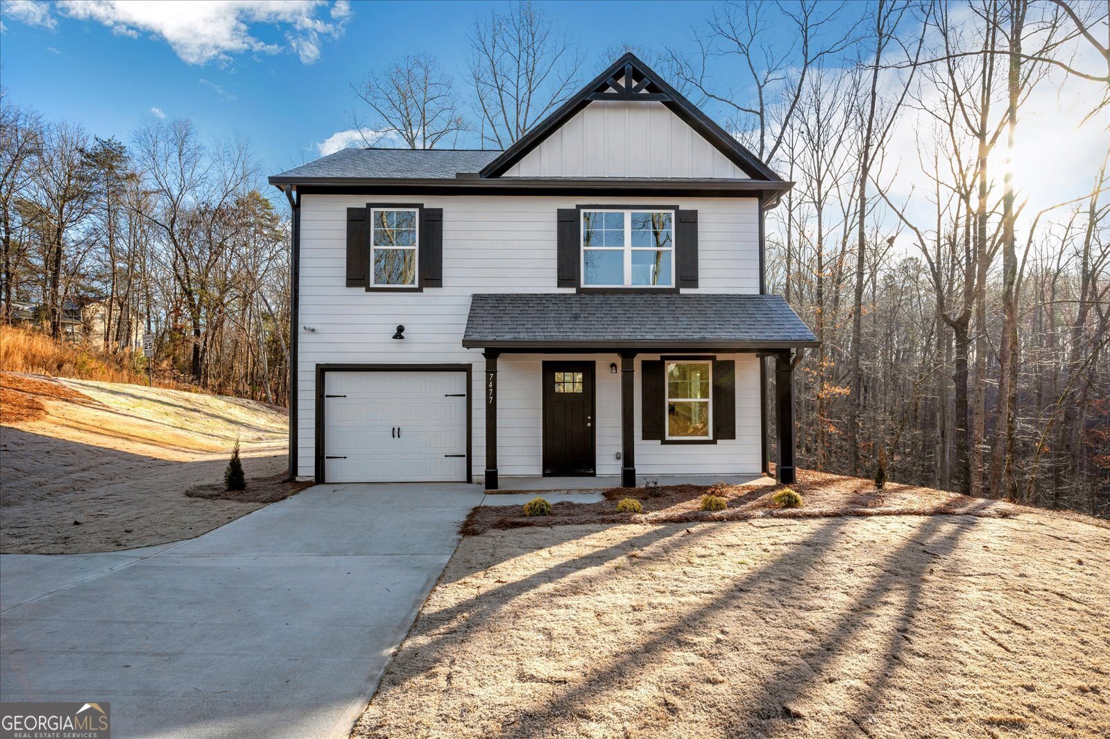 Two-story white house with black trim, an attached garage, and a driveway bordered by leafless woods.
