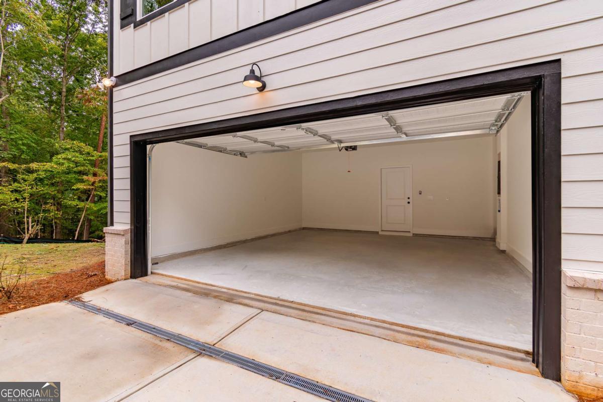 Open empty garage with a wide overhead door, bare concrete floor, and wooded yard beside the house.