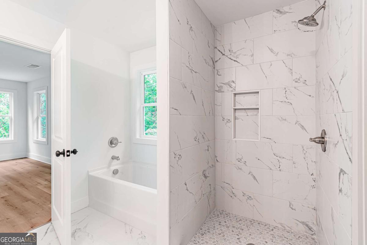Bathroom with marble-look tiled walk-in shower and bathtub beside a wood-floored room.
