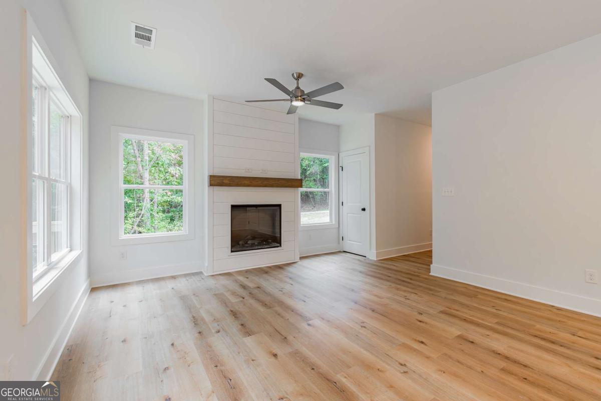 Empty living room with a shiplap fireplace, wood mantel, light wood floors, and windows overlooking trees.