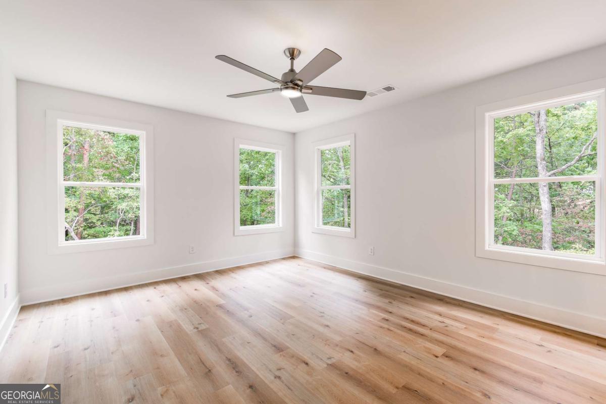 Empty room with hardwood floors, a ceiling fan, and windows overlooking trees.