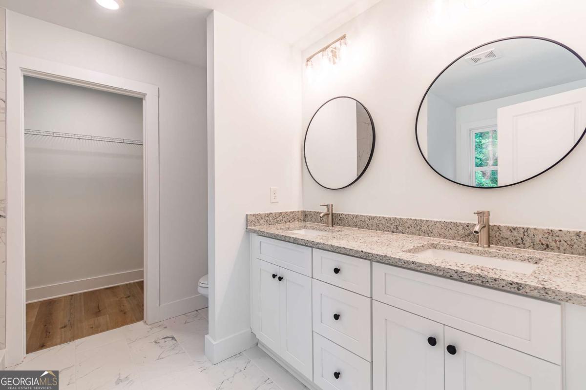 Bathroom with a white double vanity, speckled stone countertop, and two round mirrors beside an open closet.