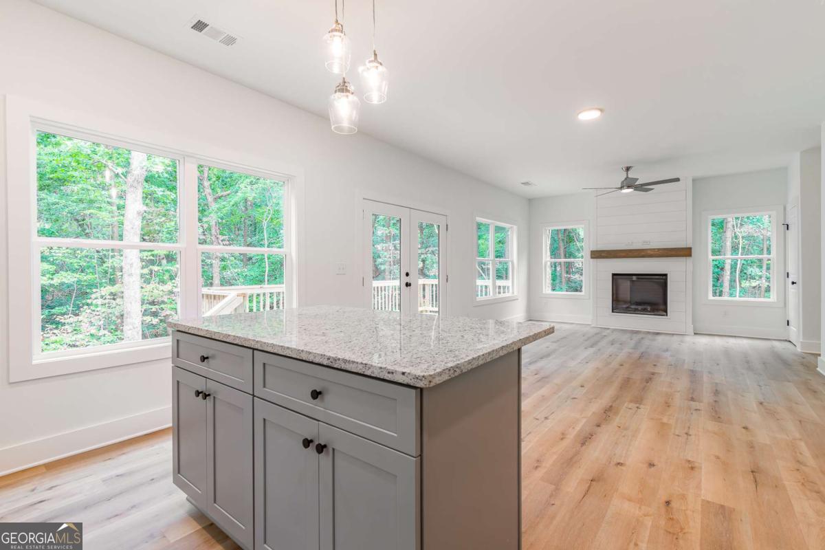Open-plan kitchen with a large gray island, light wood floors, and a fireplace beside wooded views.