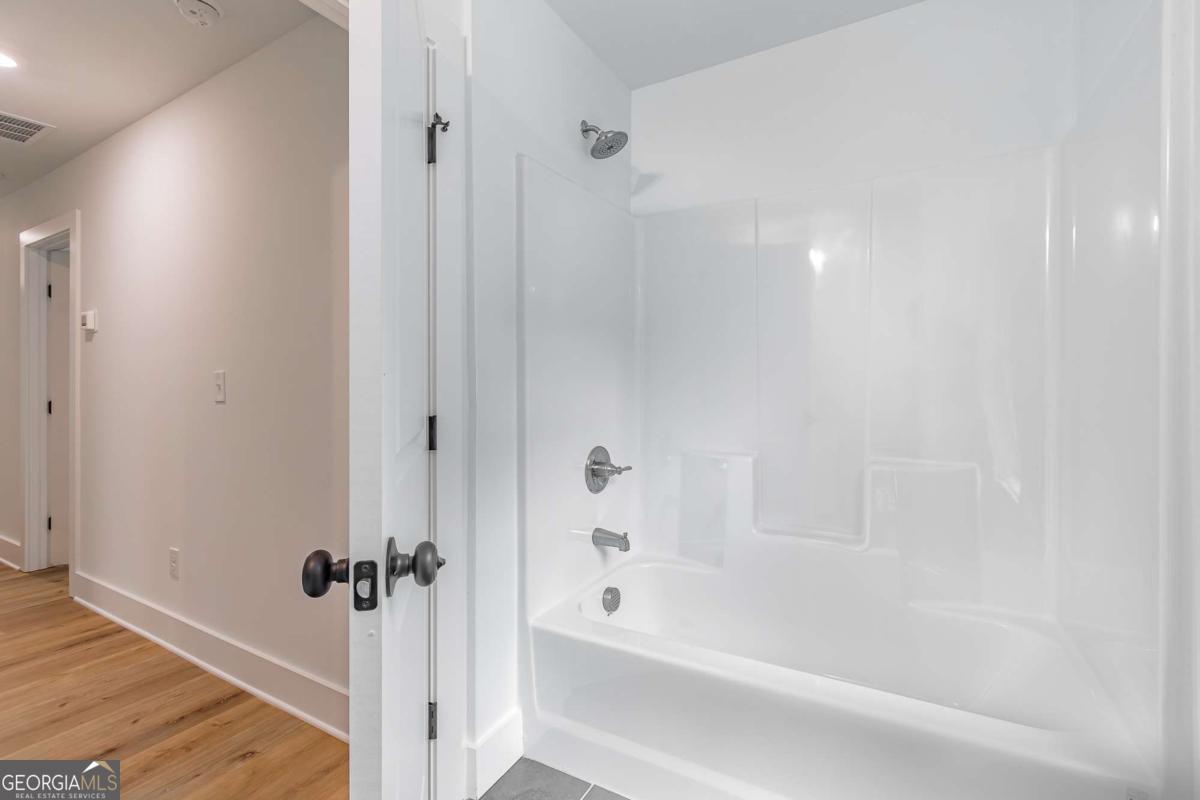 Bathroom with a white tub-and-shower enclosure and chrome fixtures beside a wood-floored hallway.