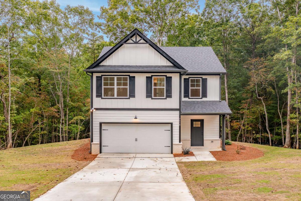 White two-story house with black trim, a front gable, and an attached two-car garage amid woods.