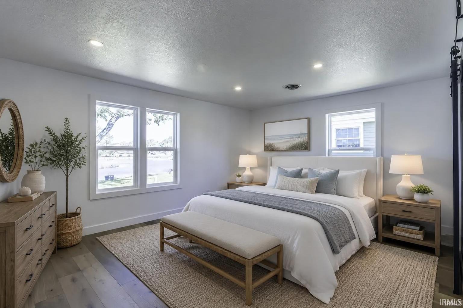 Staged bedroom with a white bed, gray bedding, a bench, and two windows.
