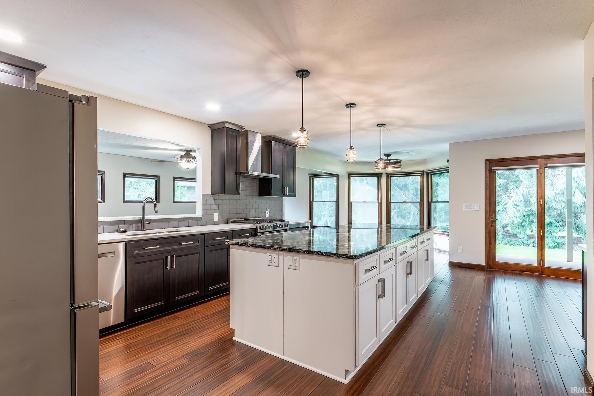 Open kitchen with a large dark stone island, dark cabinets, and windows beside glass doors.