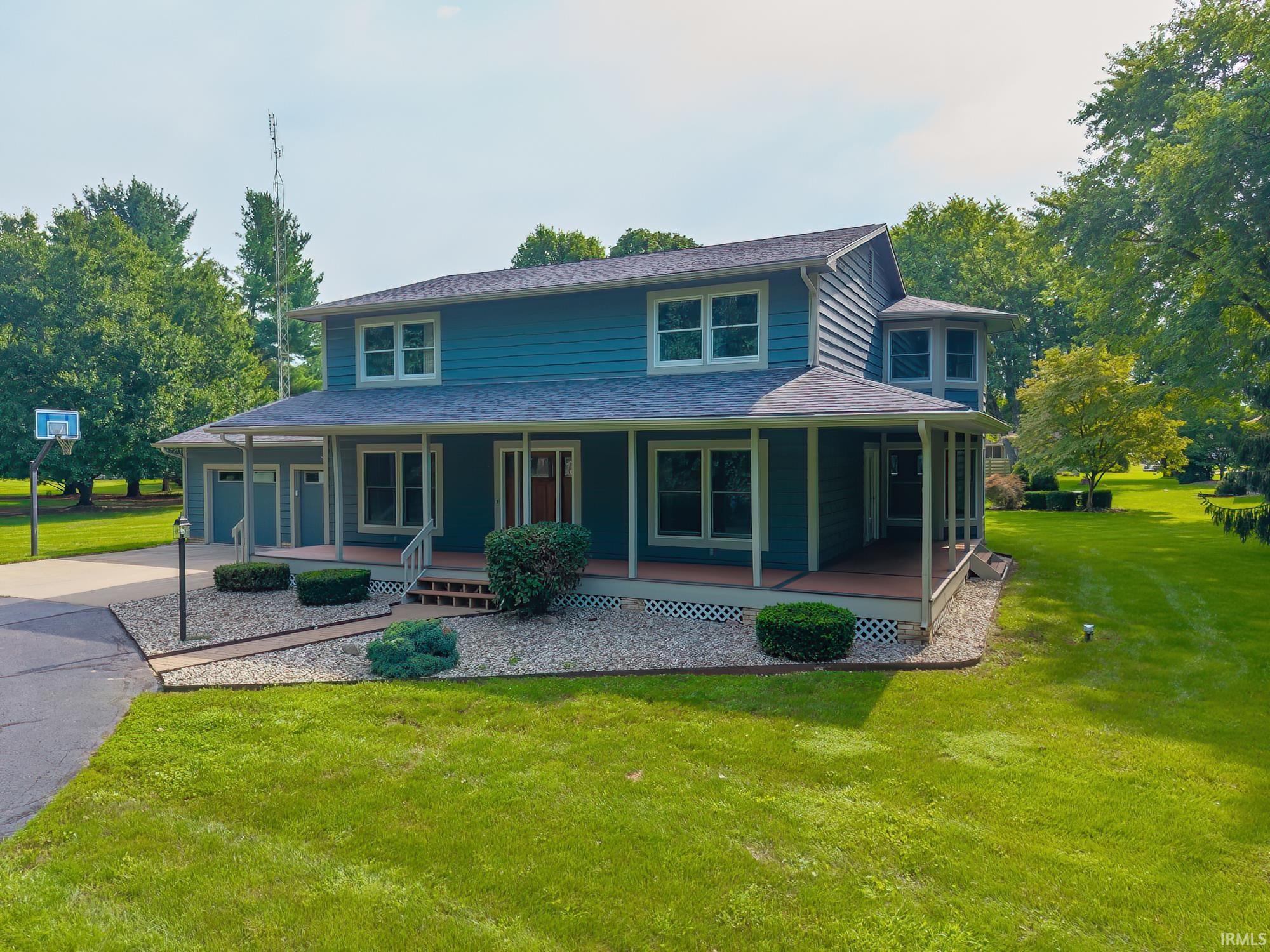 Blue two-story house with a covered wraparound porch, gravel landscaping, and a basketball hoop in the yard.