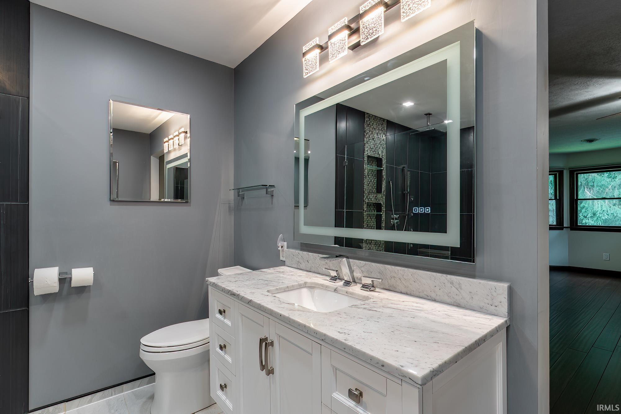 Modern bathroom with a marble vanity, illuminated mirror, and dark tiled shower reflected in the glass.