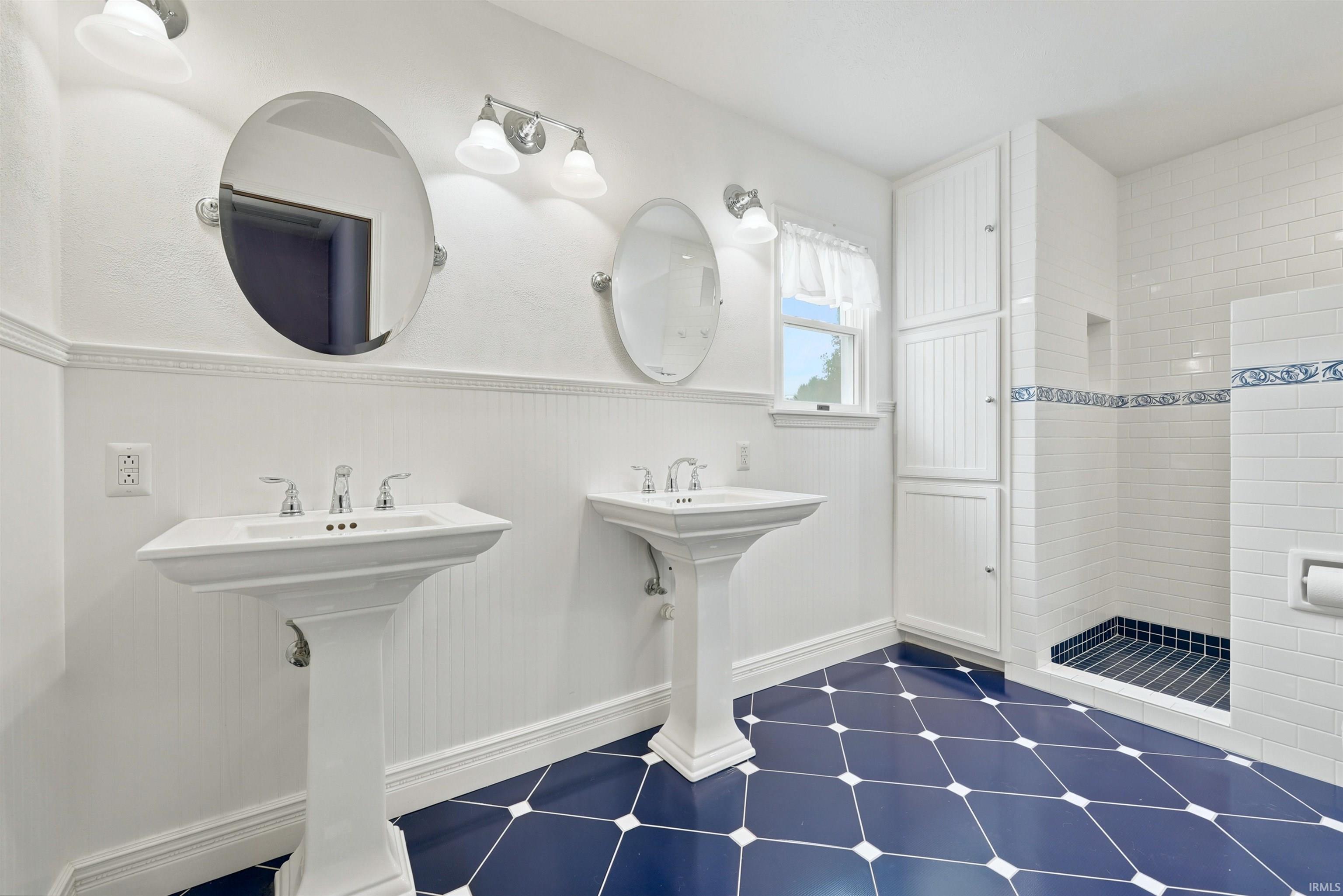 Bathroom with two pedestal sinks, round mirrors, white tile, and a dark blue geometric floor.