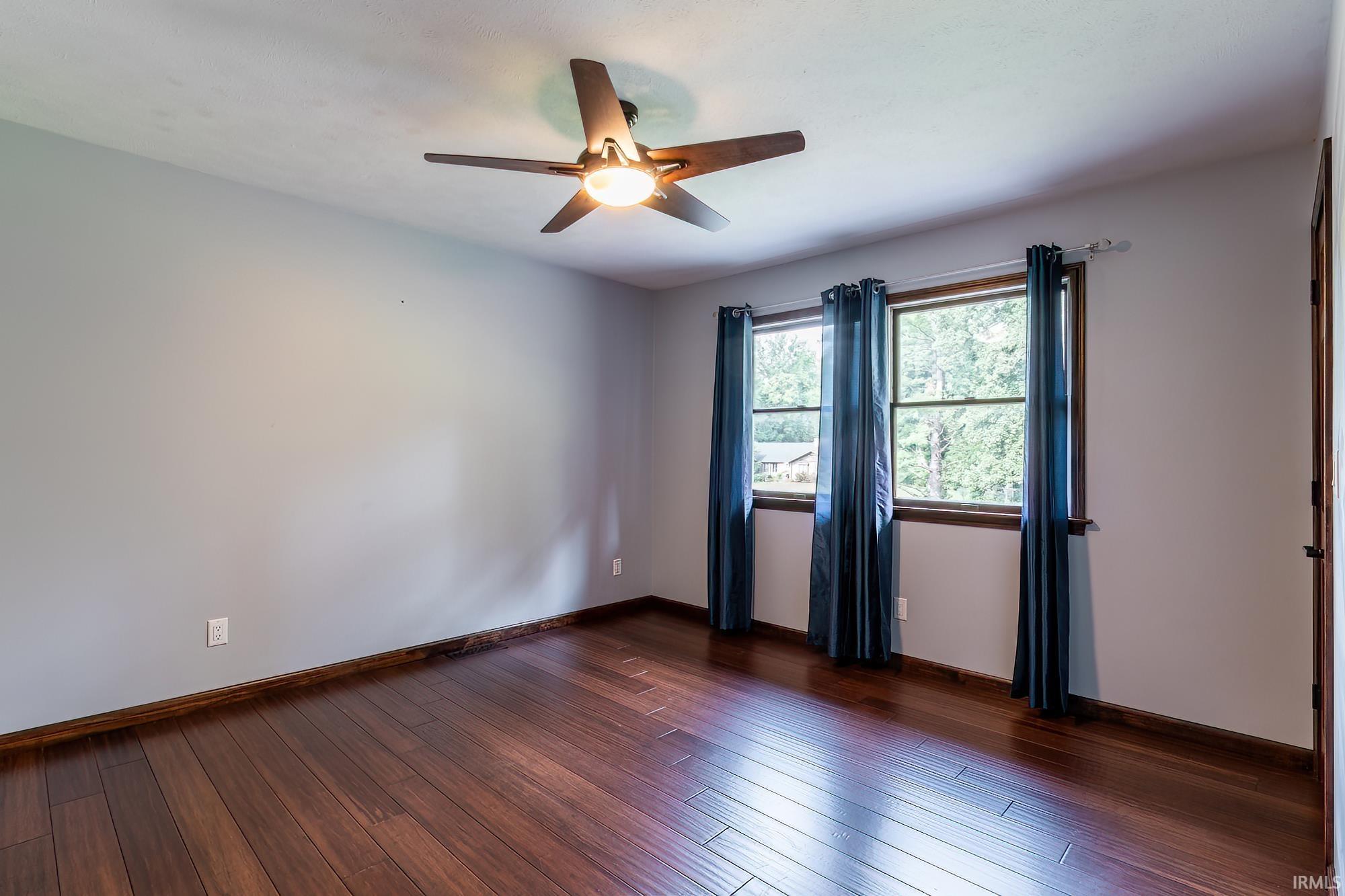 Empty bedroom with dark wood flooring, blue-curtained windows, and a ceiling fan.