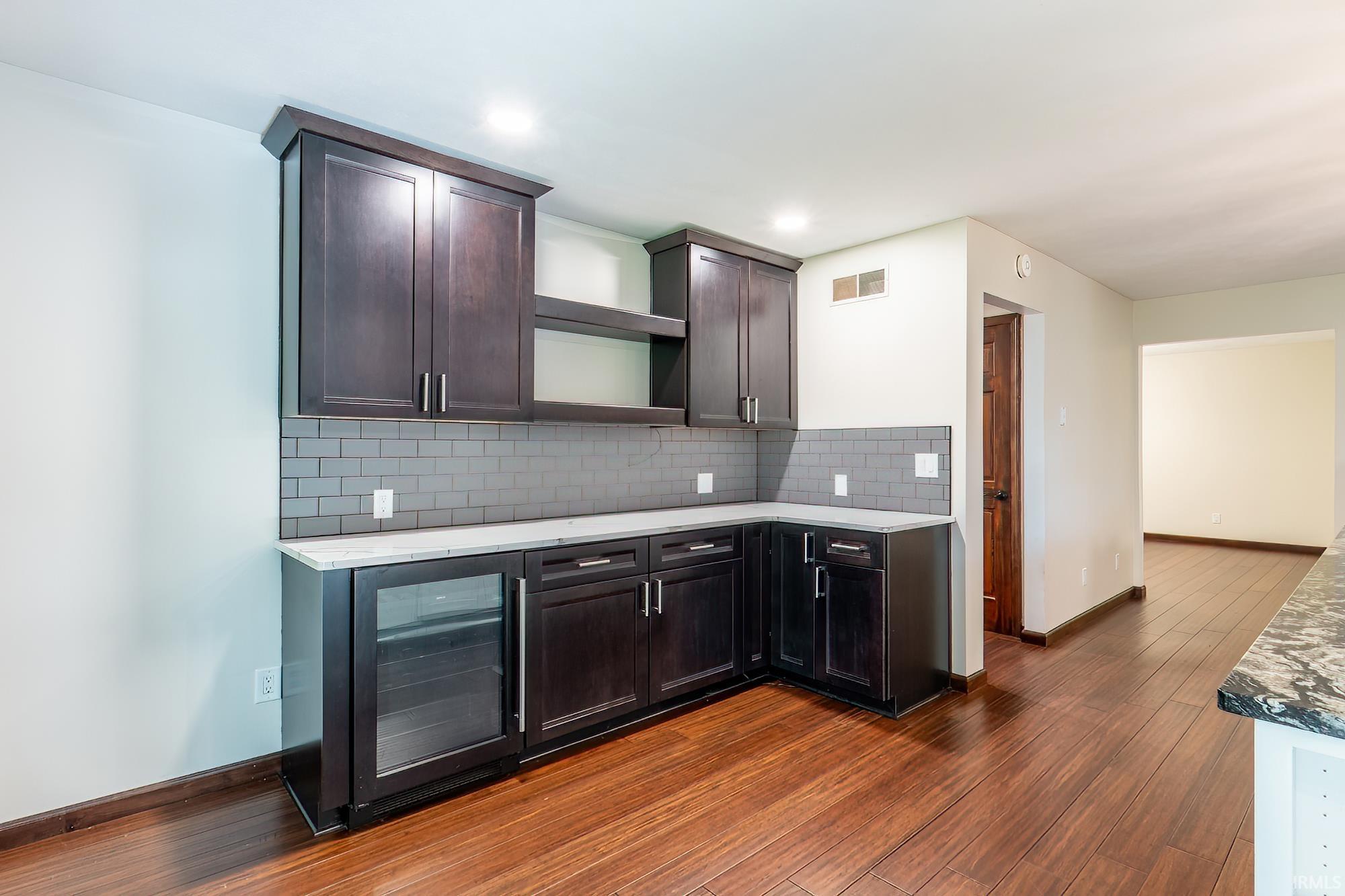 Kitchen with dark cabinets, gray tile backsplash, white counters, and warm wood flooring.