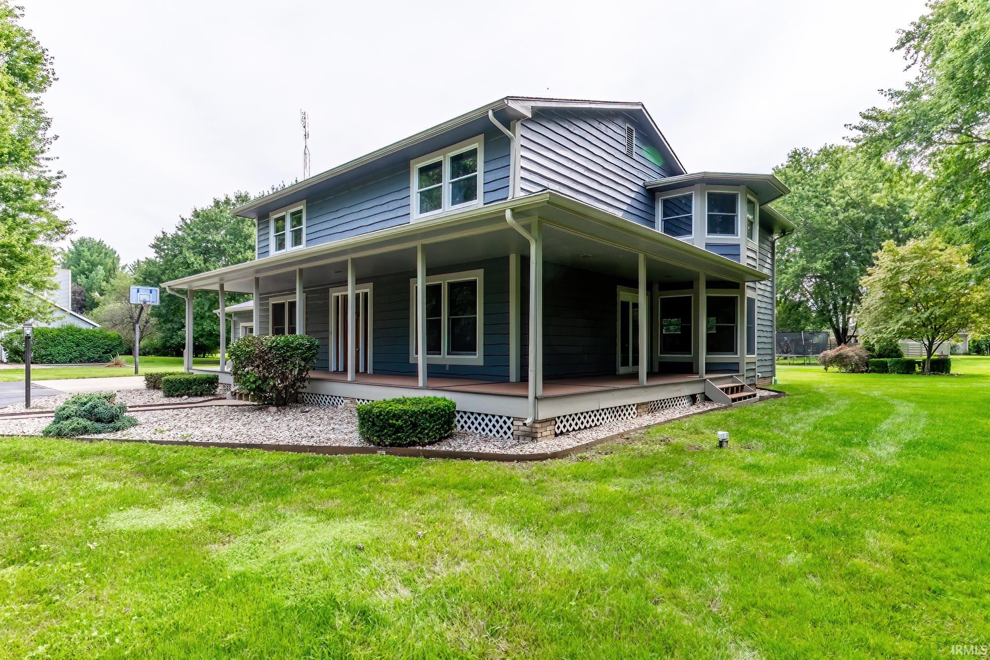 Two-story blue house with a covered wraparound porch, landscaped beds, and a broad green lawn.