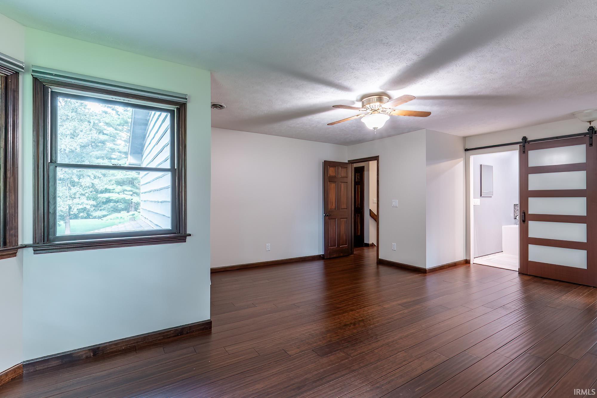 Empty room with dark hardwood floors, a ceiling fan, large window, and sliding barn-style door.